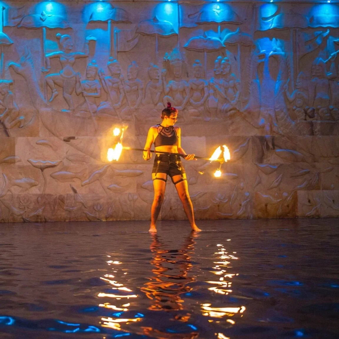 A woman performing a fire dance in front of a carved stone wall, with reflections on water.
