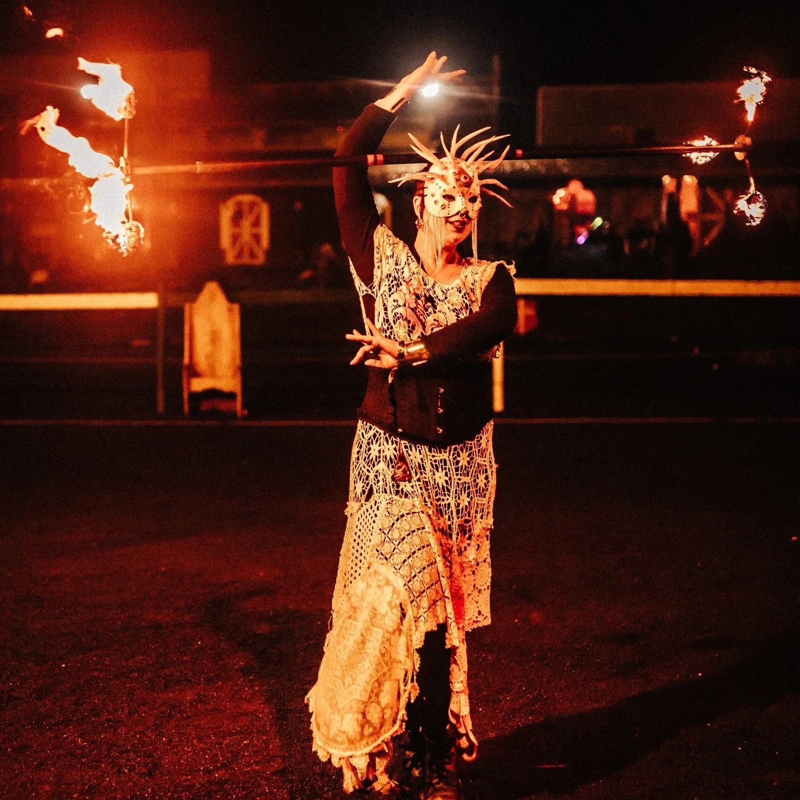 A performer dressed in a costume with lace, black clothing, and an elaborate mask with spikes, performing a fire dance at night.