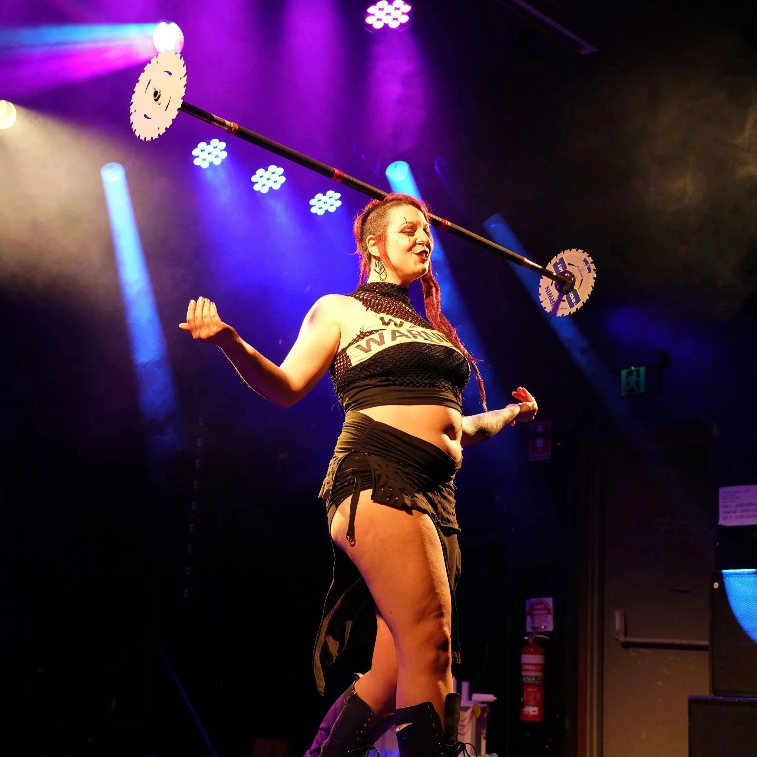 Performer on stage balancing a long pole with circular saw blades on each end, wearing a black crop top and shorts, with colorful stage lighting.