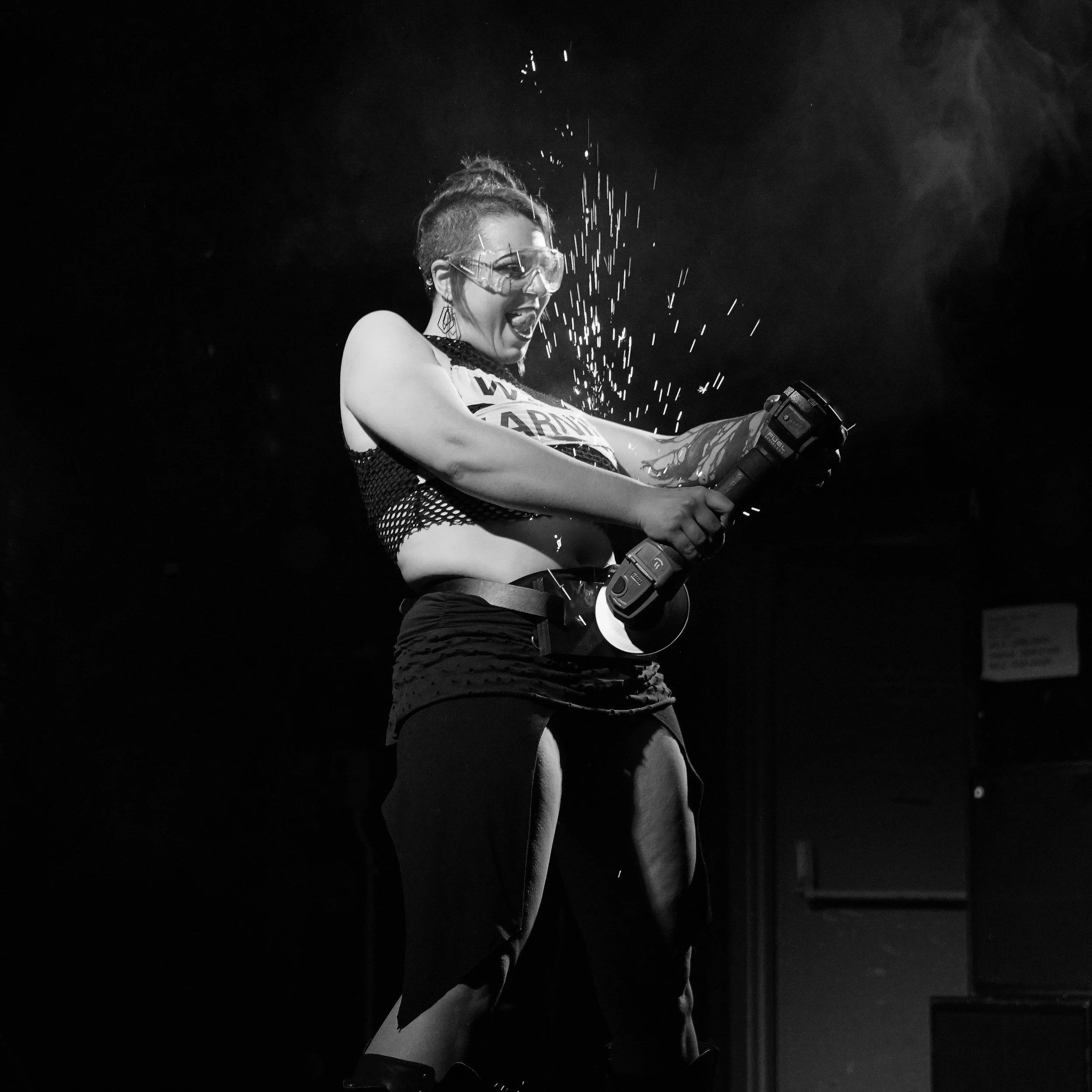 A person in safety glasses and punk style clothing is joyfully using a power drill, with sparks flying around, in a dark setting.