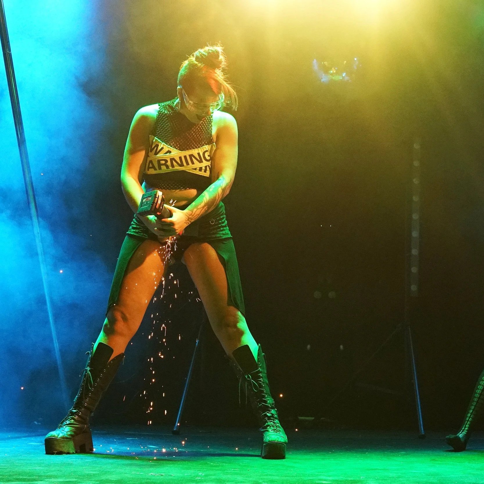 A person with tattoos wearing a black, sleeveless top with a 'Warning' sign, black shorts, and tall black boots is standing on a stage with colorful lighting. The person is holding a power tool, sparks are flying, and they are wearing glasses.