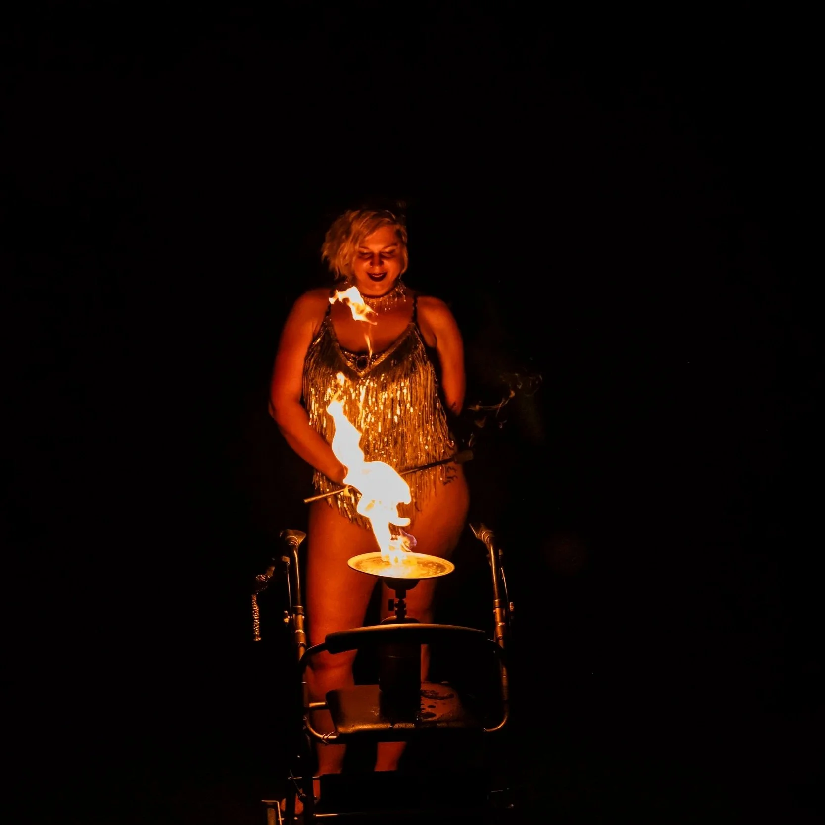 A woman in a sparkly gold dress performing fire-eating with flames on a torch in a dark setting.
