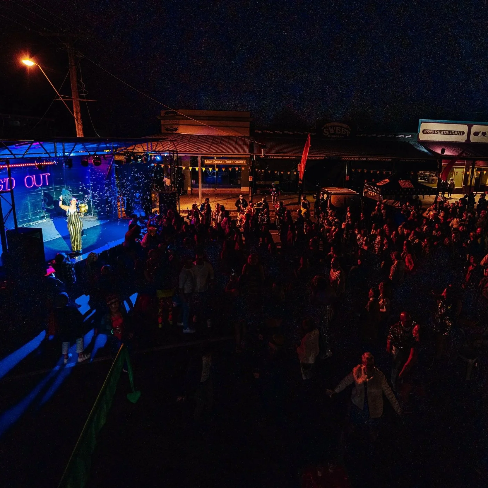 Night scene of a crowd gathered at an outdoor event with a small stage where a performer is singing, surrounded by colorful lights, with a dark sky and stars visible.