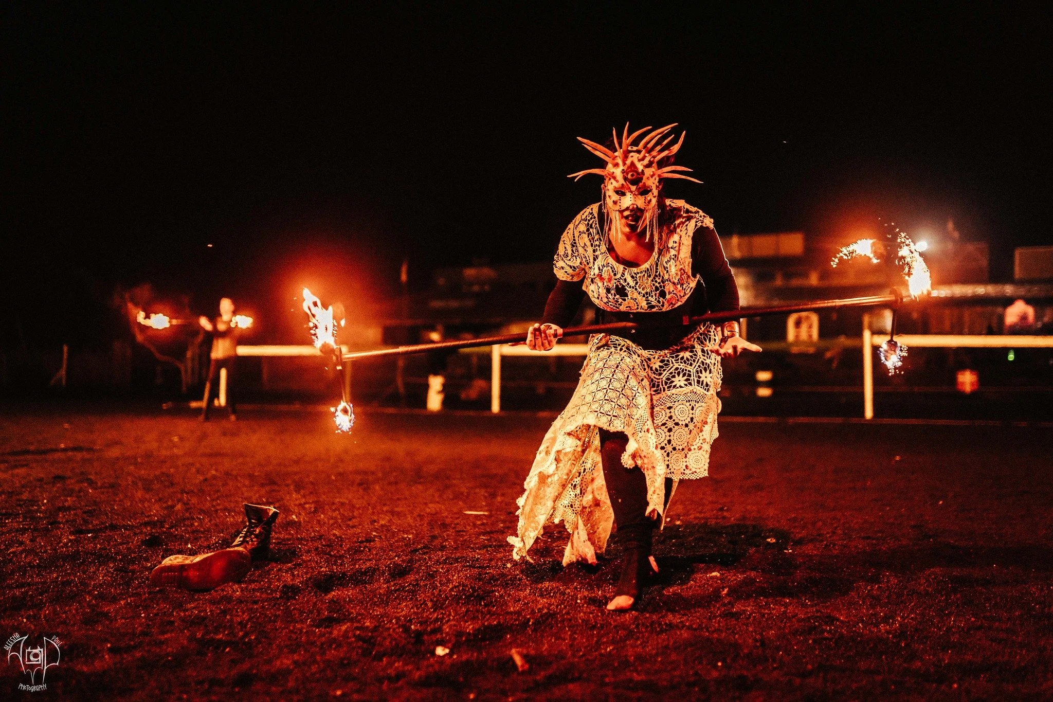 Person wearing an elaborate mask with spikes and designs, dressed in lace clothing, performing a fire dancing routine at night on a field.