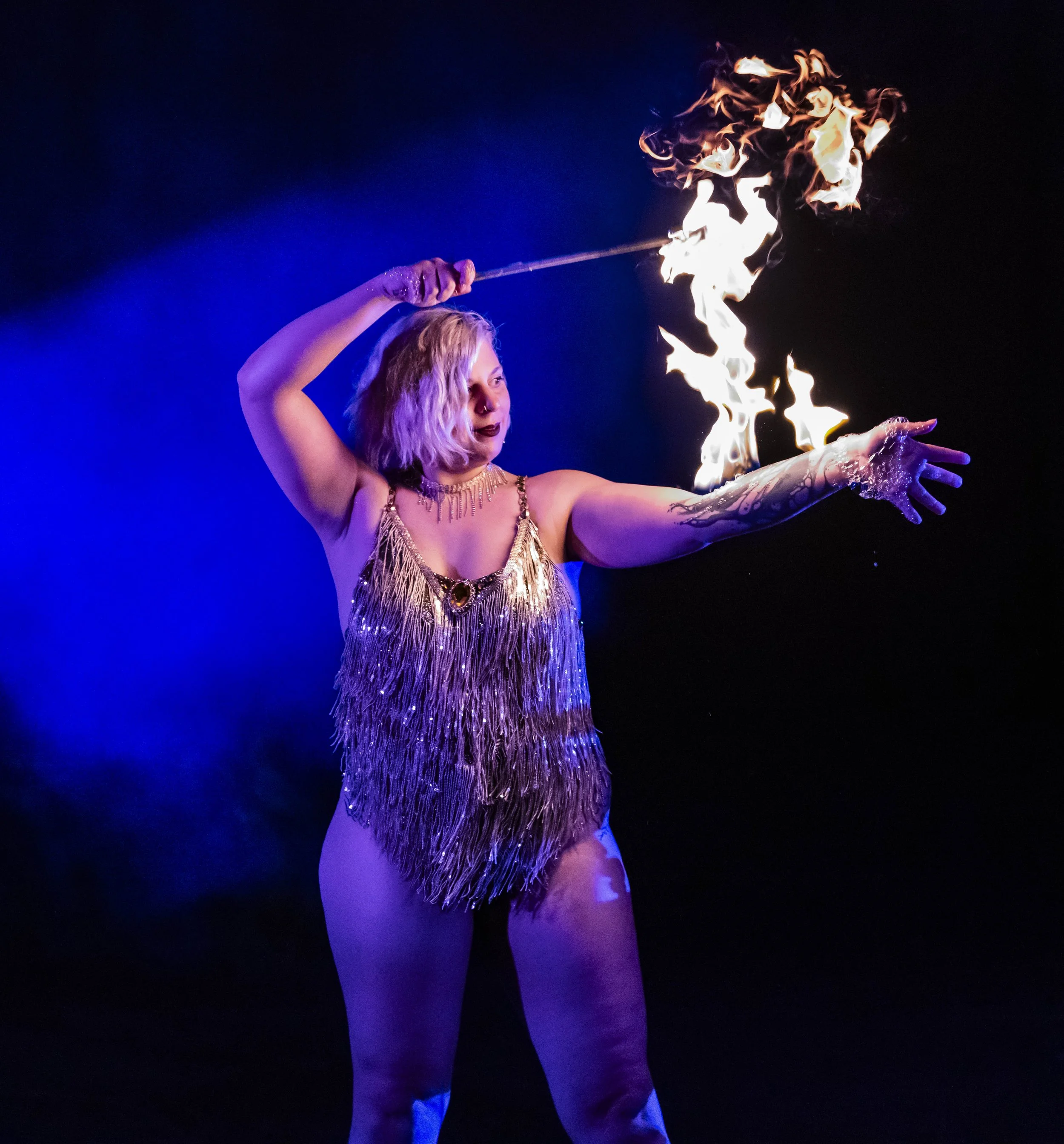 A female performer in a fringed gold outfit with a tattooed arm, holding a flaming torch, performing a fire act on stage with dark background and blue lighting.