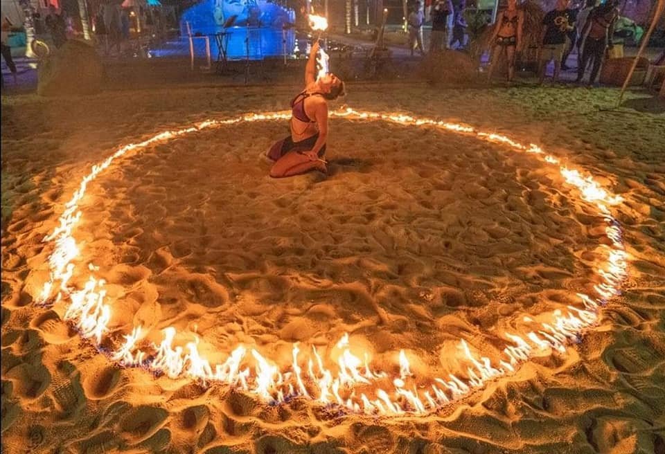 A woman performing a fire dance inside a circle of fire on a sandy beach at night during a festival or event.