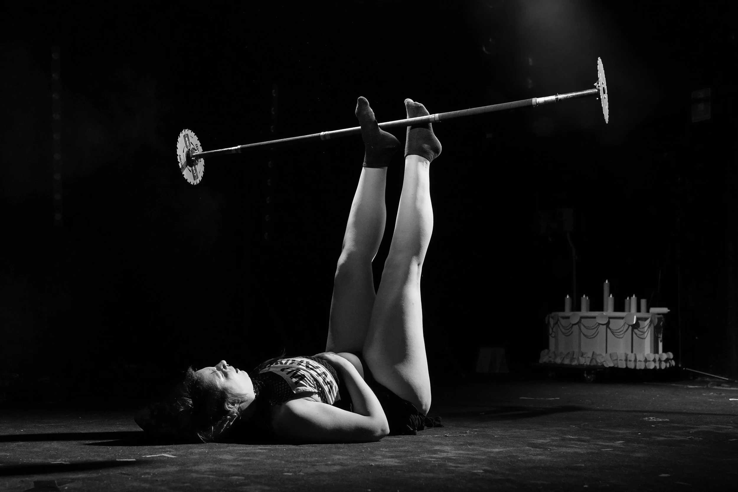 A woman lying on her back on stage, holding a barbell with two wheels above her legs, performing a fitness pose in a dark room with a cake in the background.