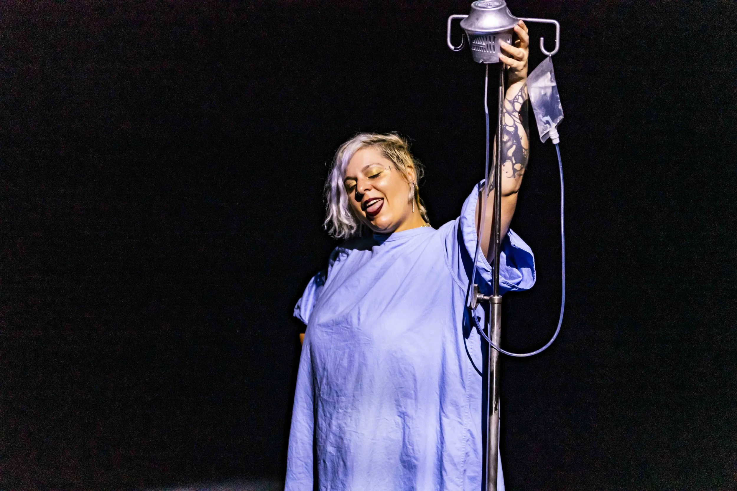 A woman with blonde hair and tattoos is in a hospital gown, holding an IV pole, with her eyes closed and tongue sticking out in a playful pose against a black background.