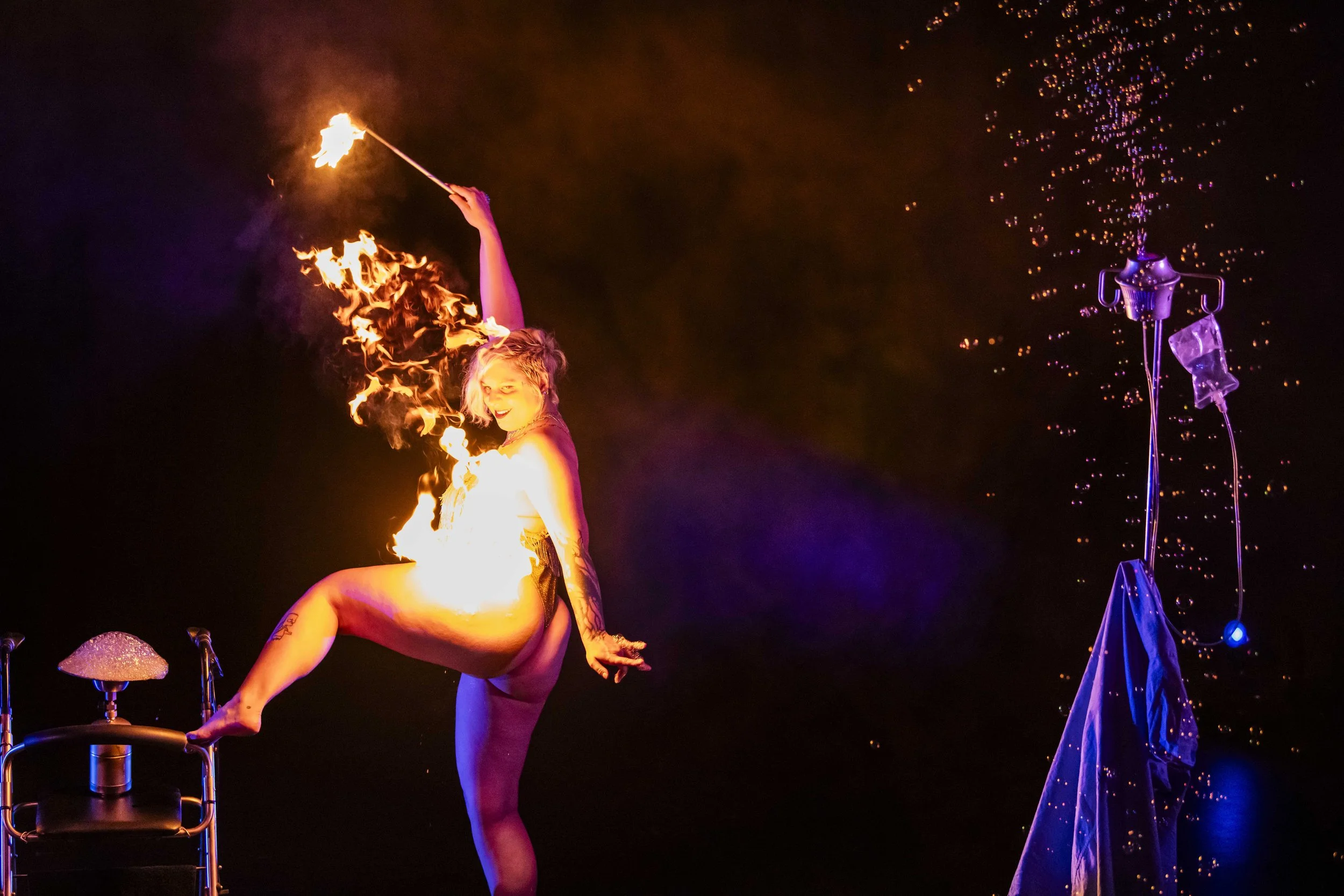 Performer with short hair and tattoos, balancing on one leg, holding a flaming torch in one hand and a fire staff in the other, with fire and sparks around her in a dark setting.
