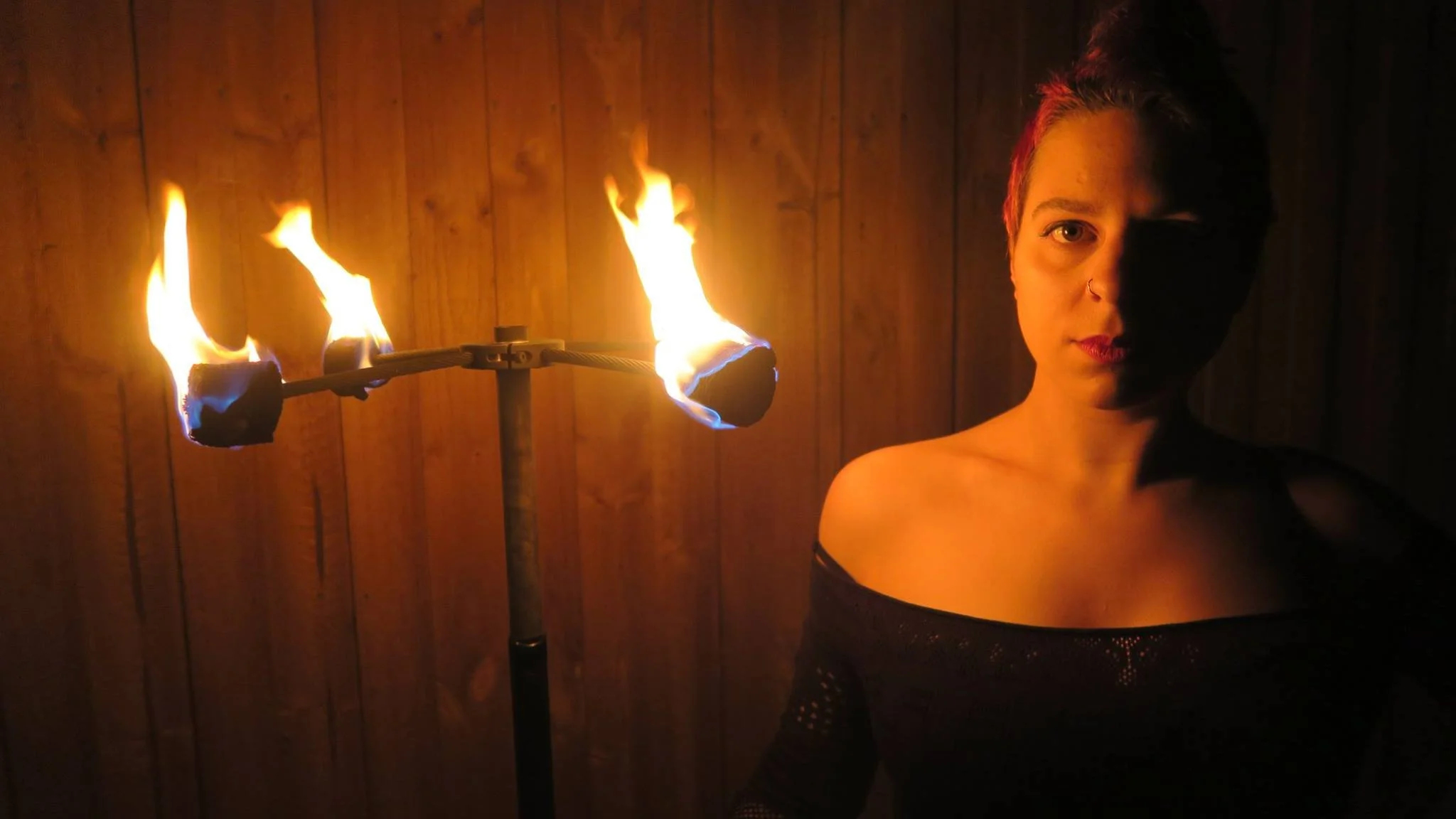A woman with short hair and a nose ring standing next to a flaming torch in a dimly lit room with wooden walls.