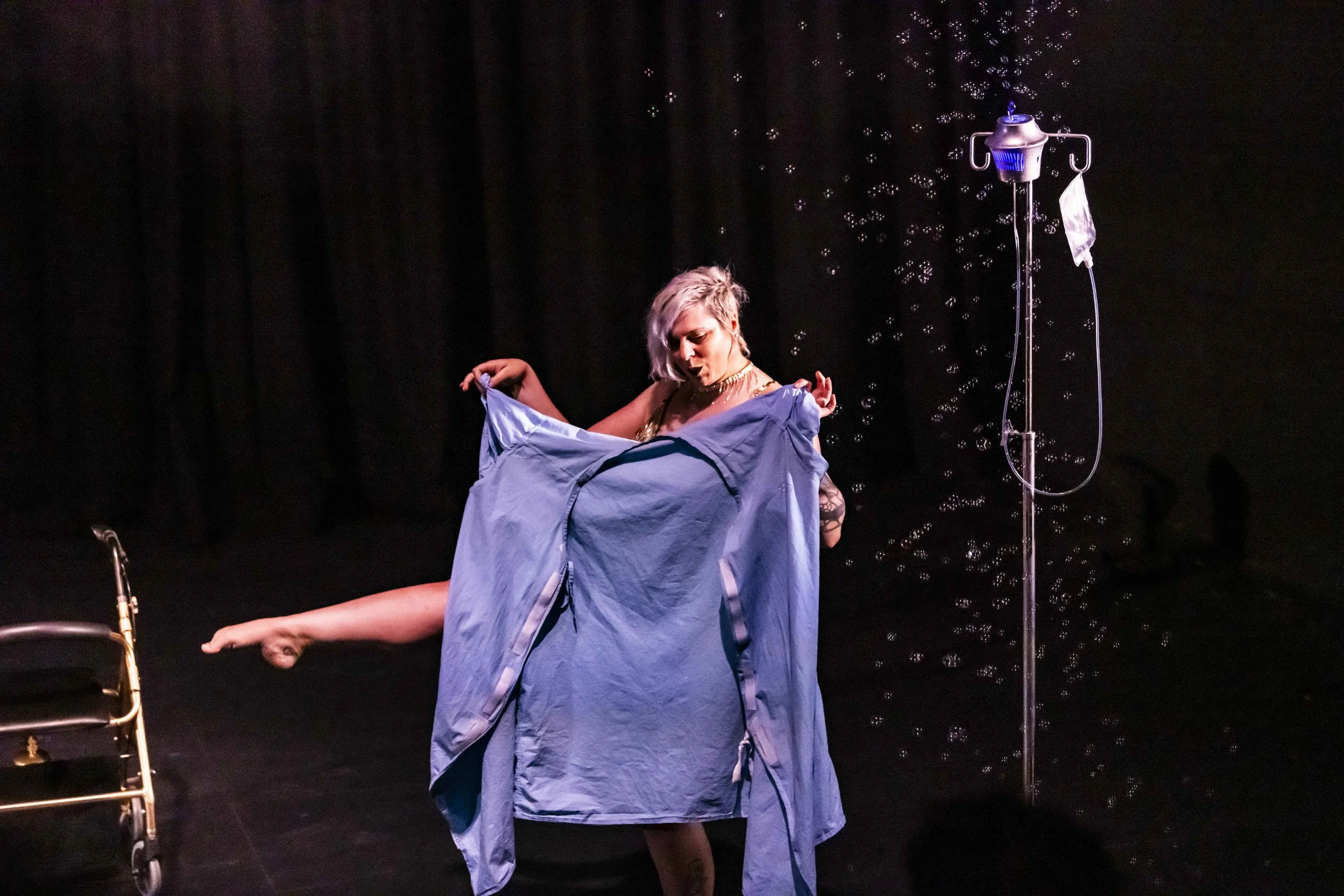 A performer on stage with short gray hair, wearing a black top and gold accessories, draping a blue hospital gown over another person's leg while standing next to an IV pole. There are bubbles around and a wheelchair visible on the left side of the s