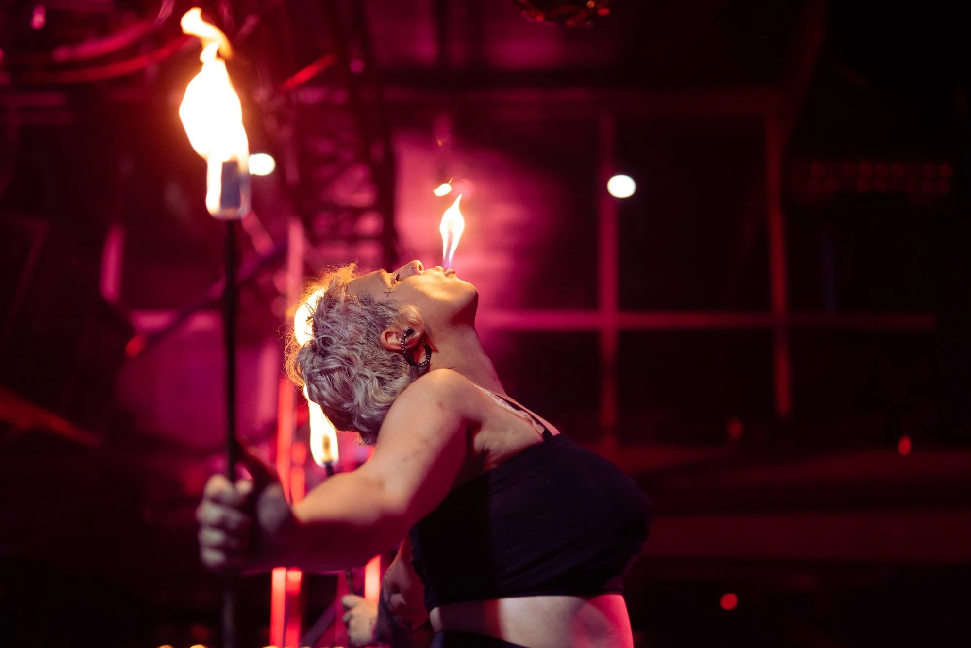 Performer with short blond hair and black outfit spinning fire poi at a dark stage with pink and red lighting.