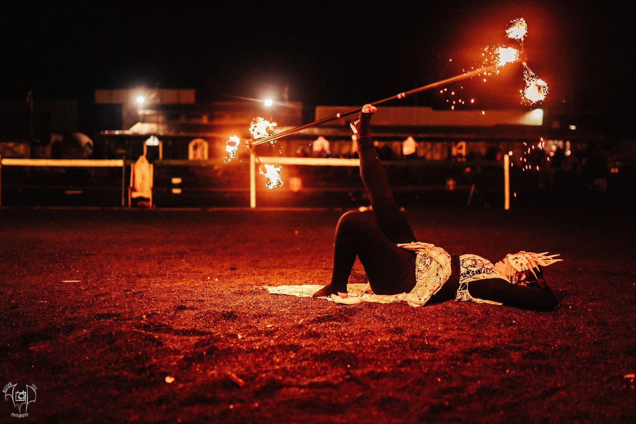A person lying on the ground at night, spinning a flaming baton in the air during a fire performance.