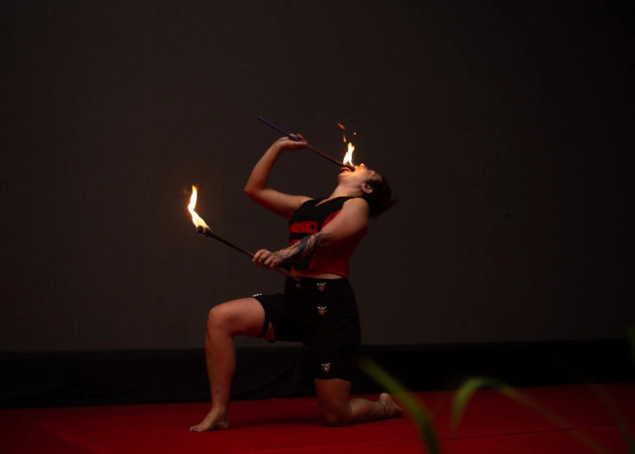 A woman performing a fire-breathing act with flaming torches, kneeling on one knee against a dark background.