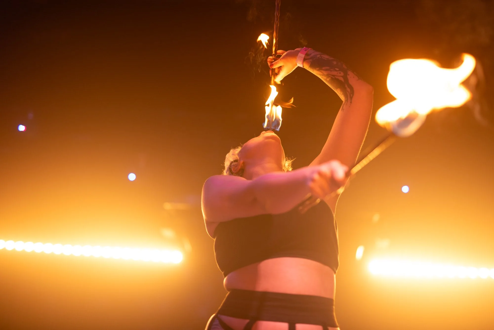 A performer spins fire on stage, tilting her head back with flames coming from her mouth and a flaming staff in her hand, illuminated by orange stage lights.
