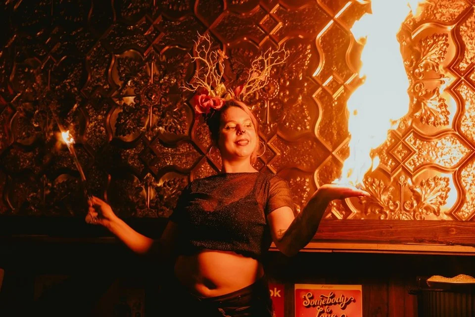 A woman performing fire with a flame coming from her hand, holding a lit torch in her left hand, in front of an ornate wall inside a nightclub or bar.
