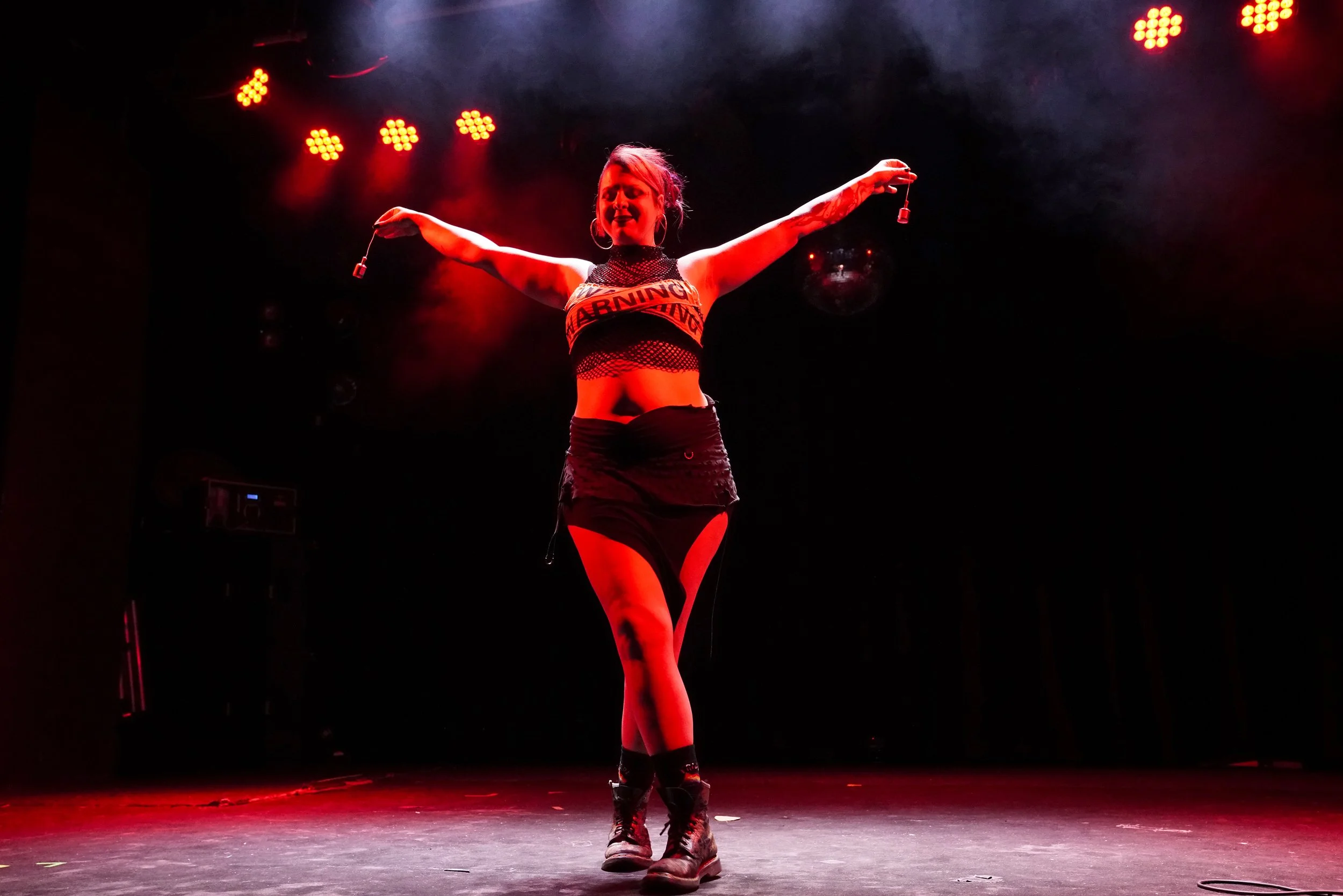 A woman performing on stage with arms outstretched, illuminated by red stage lights, wearing a cut-out black top, shorts, and black boots.