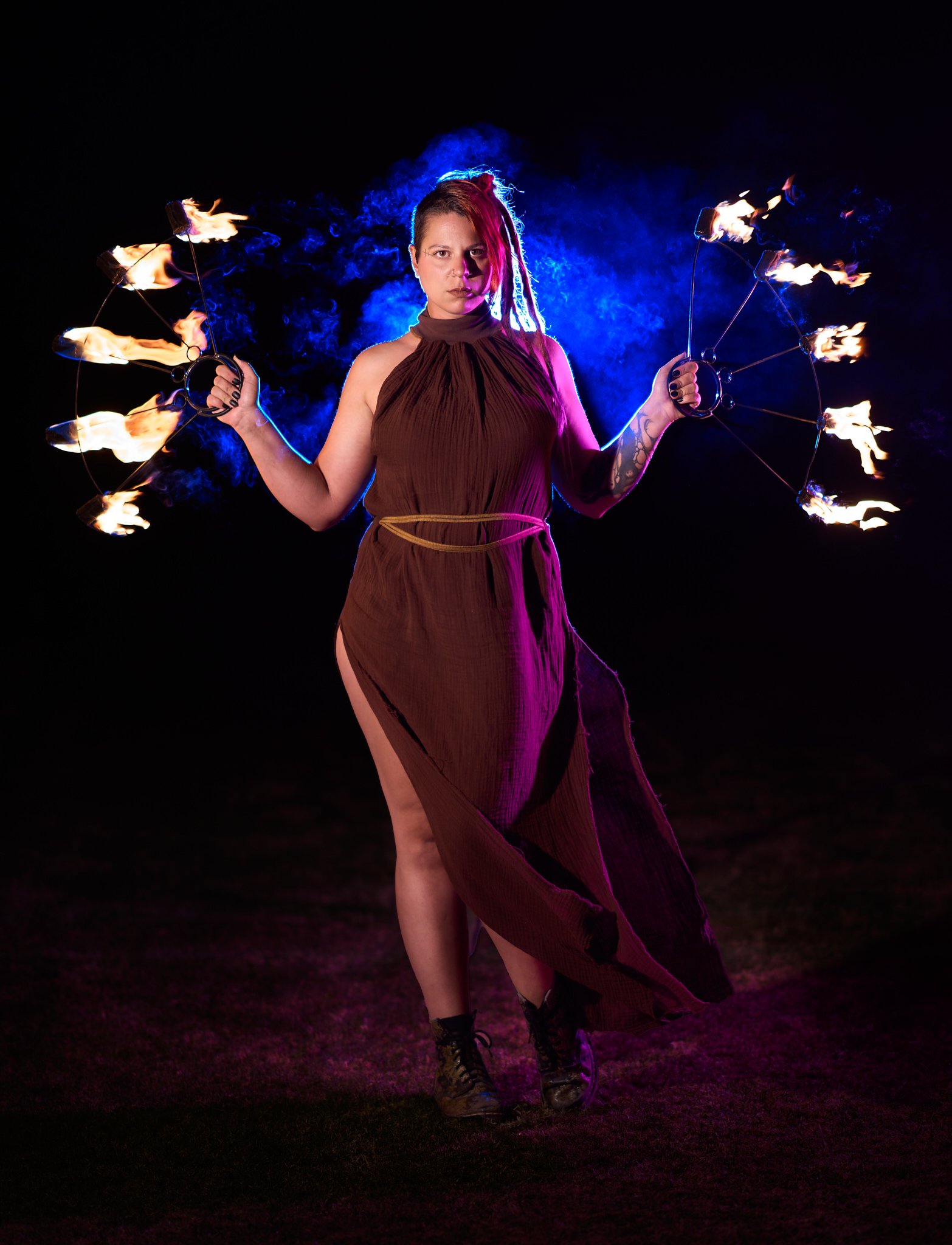 A woman with dreadlocks holds two flaming fans in a dark setting, illuminated by colorful lighting.