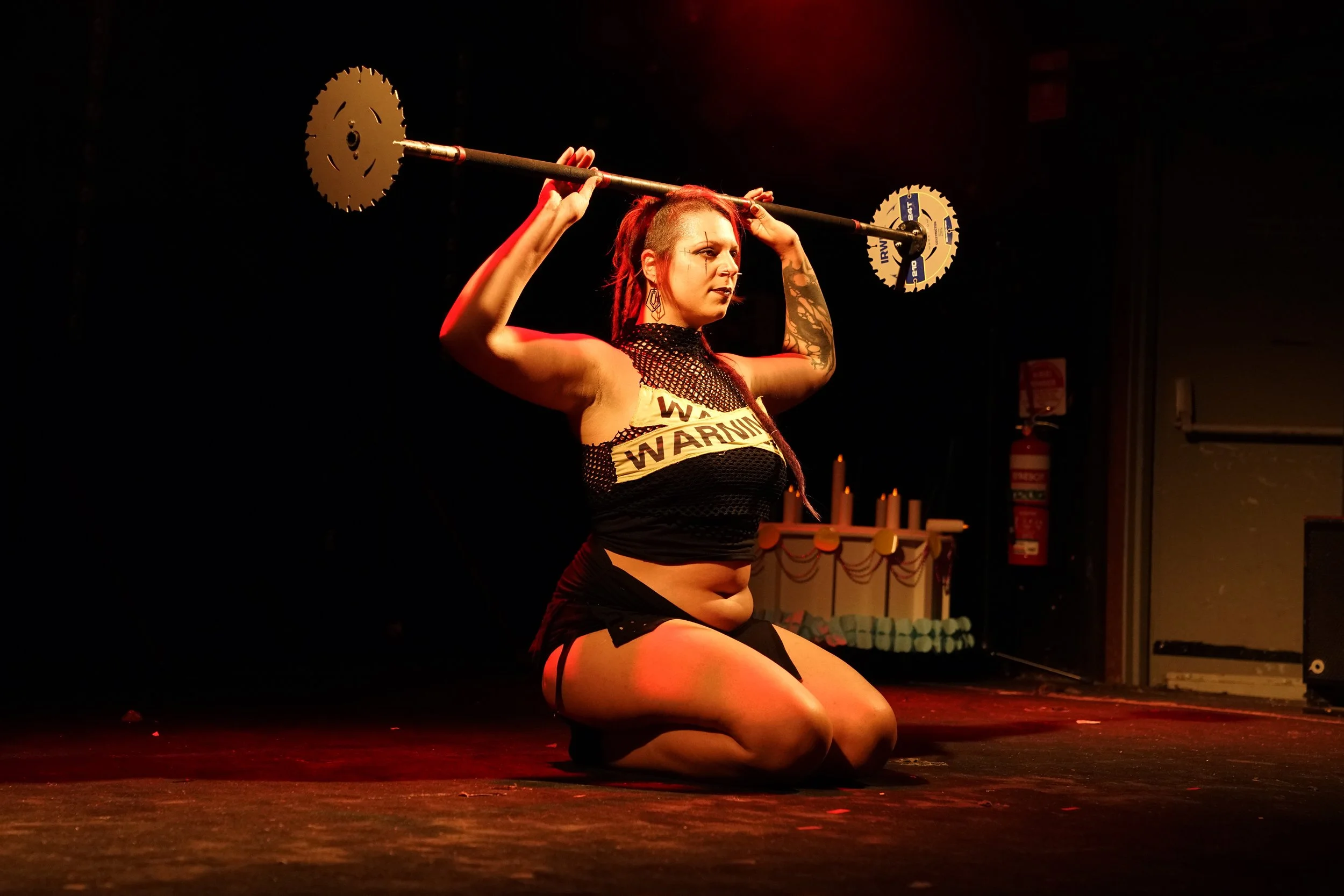 A woman kneeling on stage, holding a pole with saw blades at each end, in a performance setting with dim red lighting.
