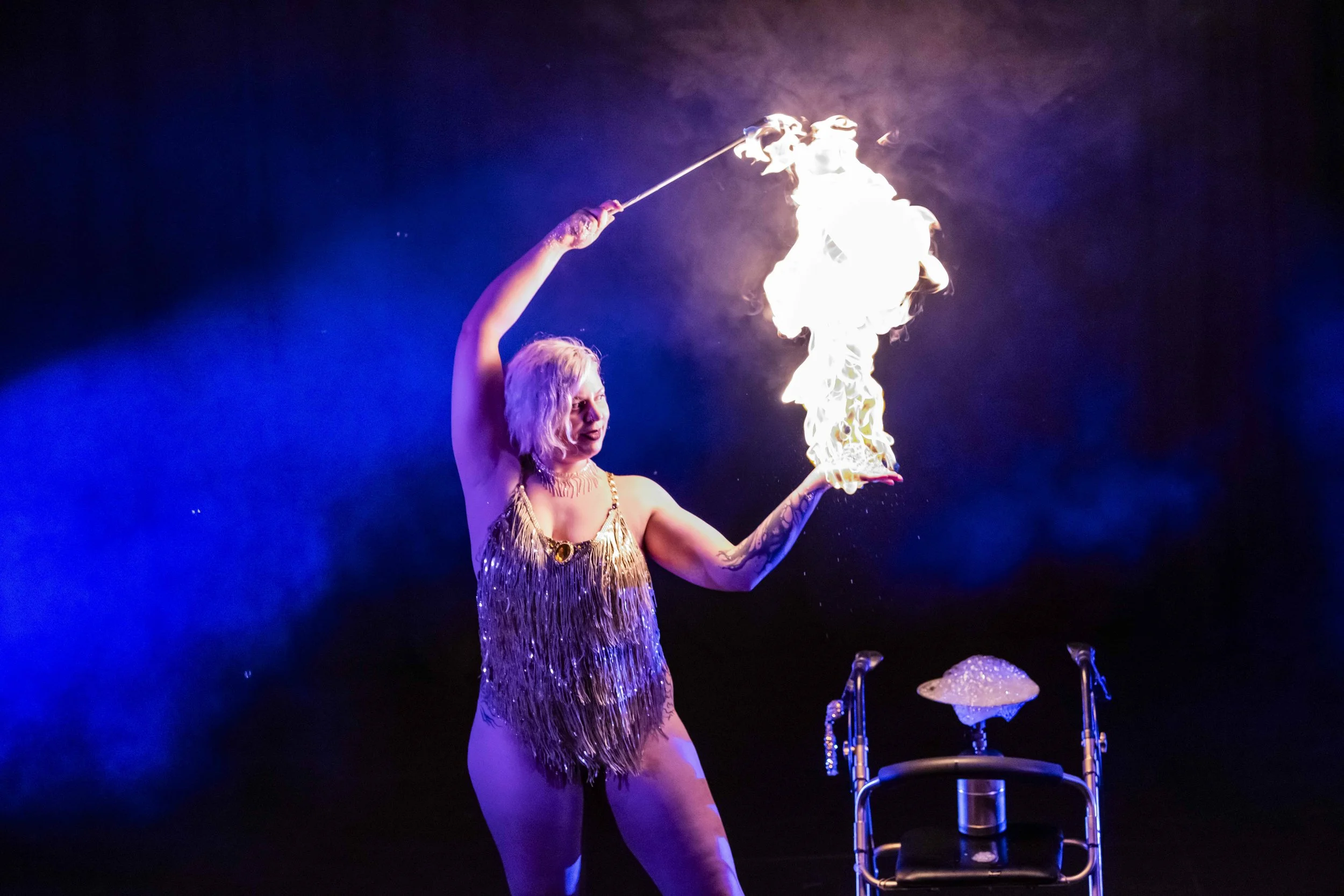 A female performer with blonde hair in a sparkly gold dress performs a fire-breathing act on stage, holding a flaming torch, with bright blue and purple stage lighting in the background.