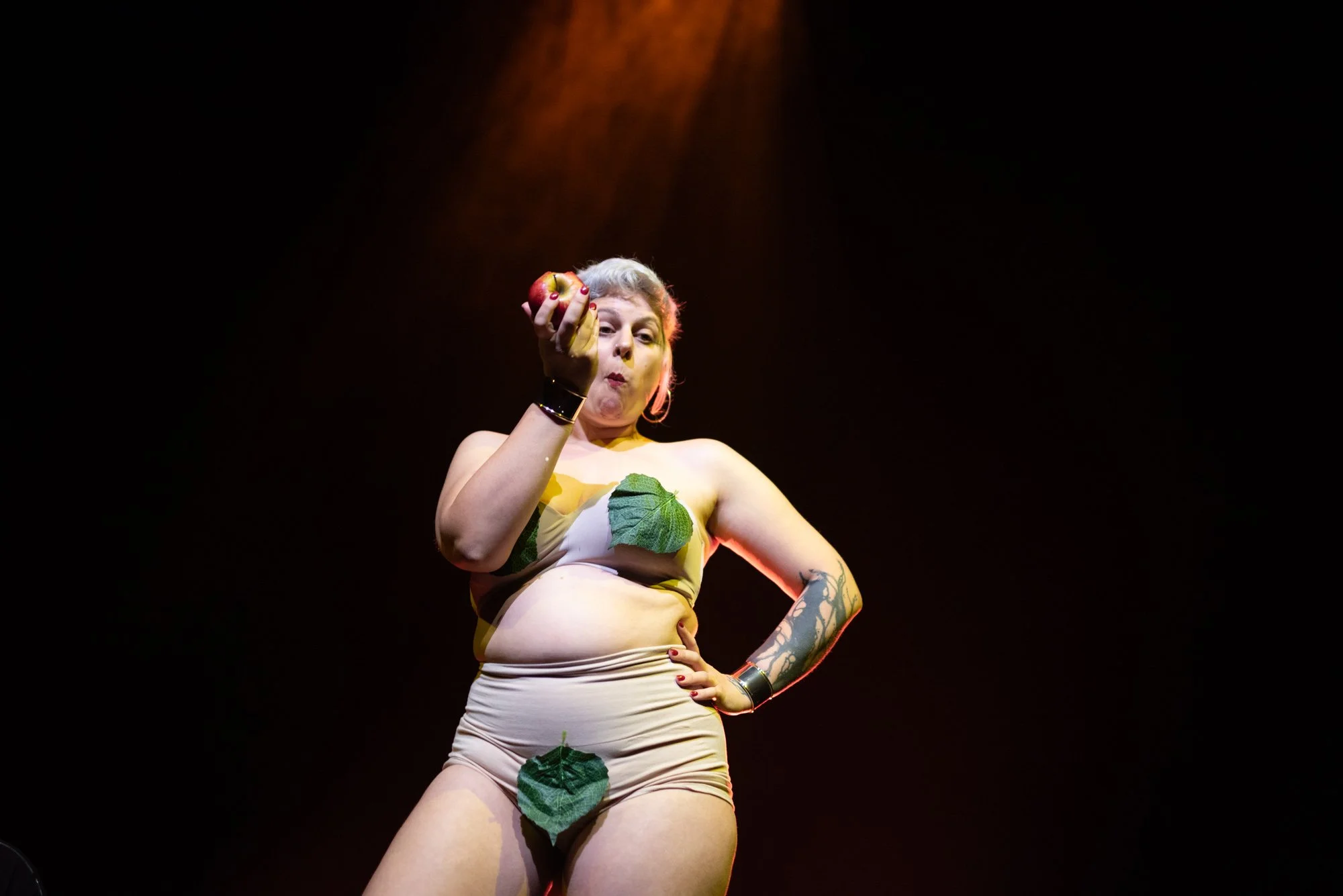 A woman standing on stage with a dark background, holding a small apple in her right hand, wearing minimal clothing covered with green leaves, and displaying tattoos on her left arm.
