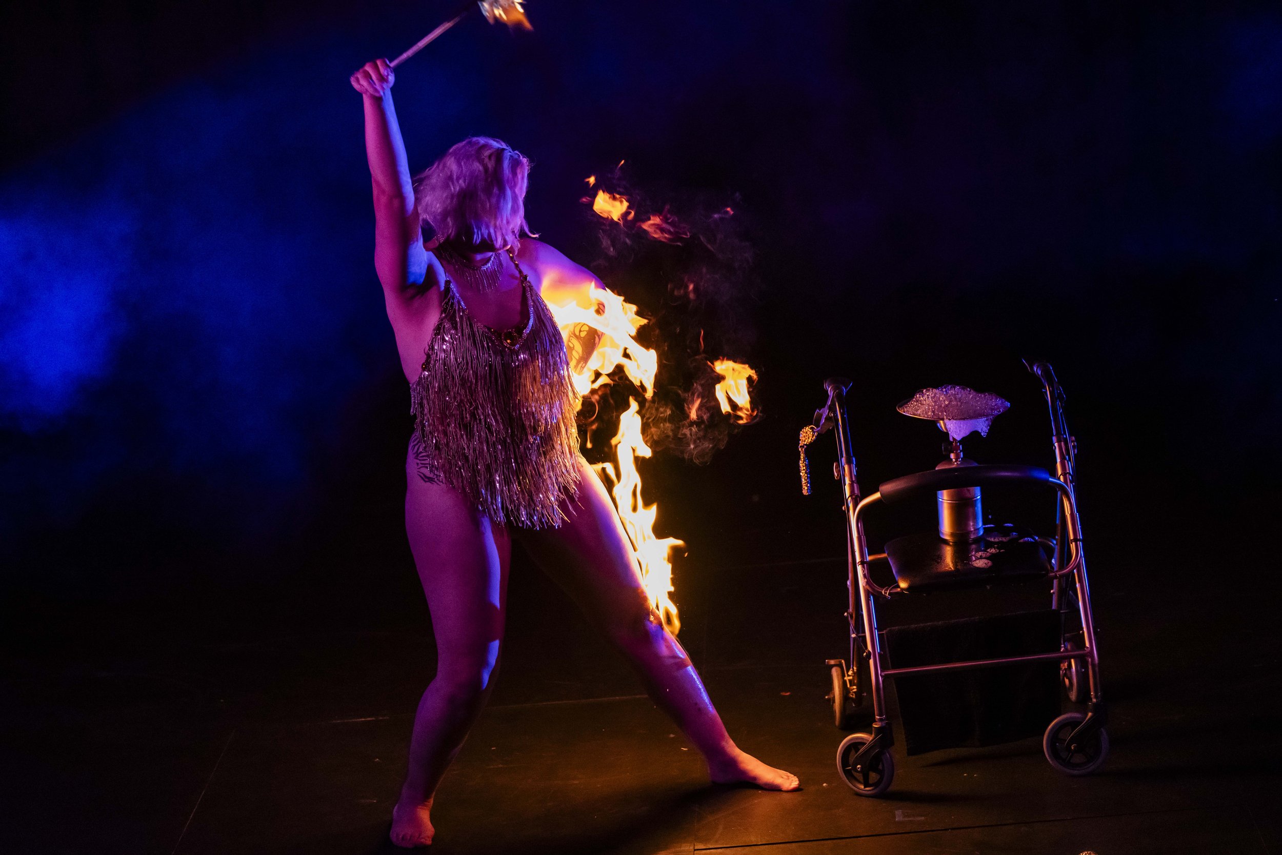 A female fire performer in a gold fringe costume performing with fire on stage, with a wheelchair nearby, against a dark background.