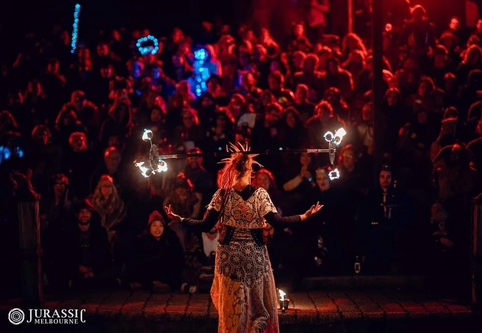 Scarab Fire balances fire prop on her head in front of a large audience at a nighttime event, with colorful lights and a dark background.