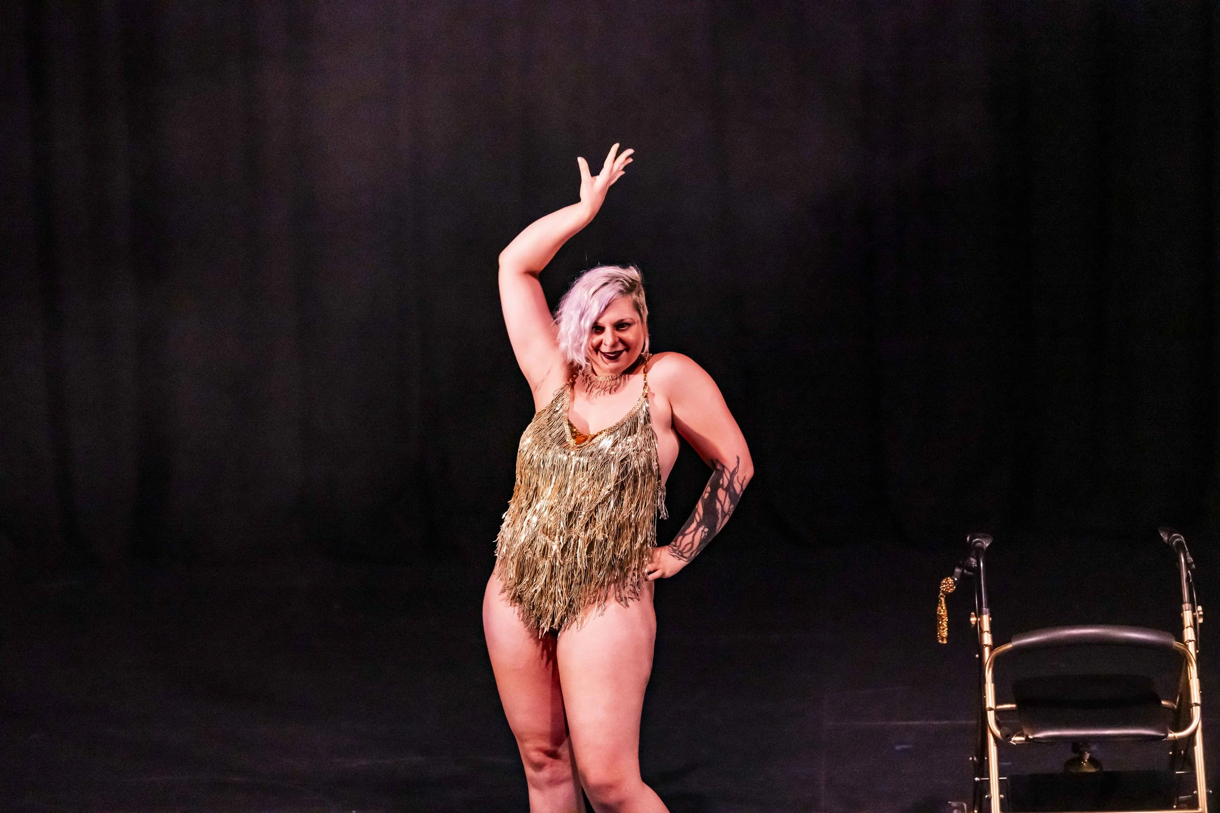 A woman with light purple hair, black lipstick, and a gold fringed dress posing on stage with a chair next to her.