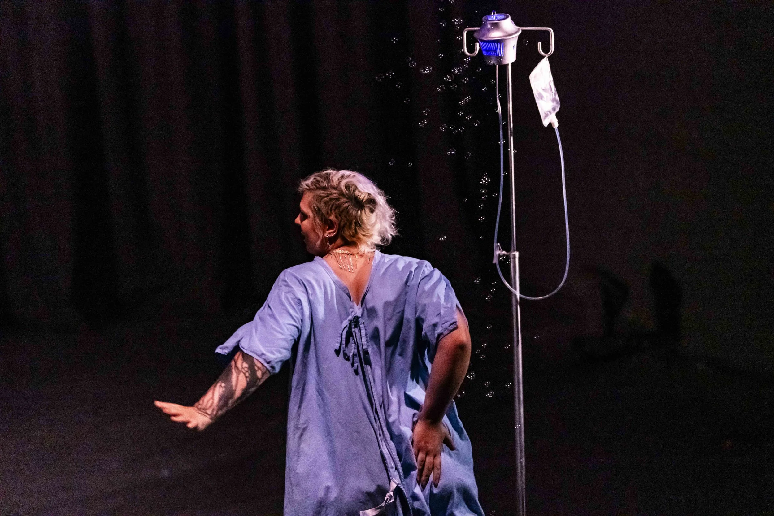 A woman in blue hospital gown performing on stage, facing away with her head turned to the side. An IV stand with a drip bag and tubing is beside her, and she is surrounded by bubbles in a dark setting.
