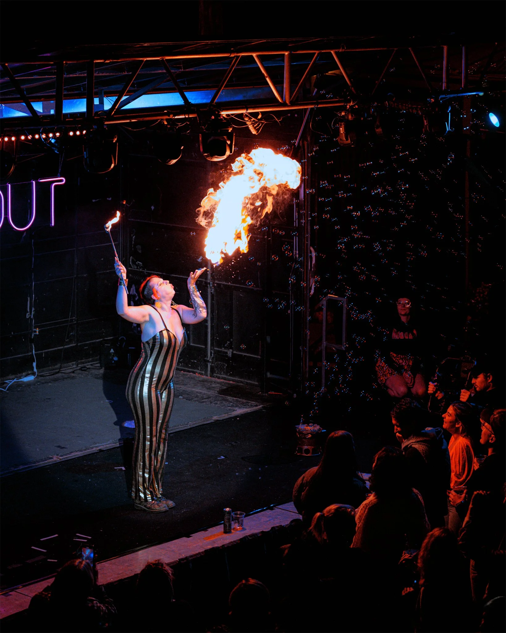 A fire performer on stage blowing fire, with an audience watching at a nightclub or theater. The stage has dramatic lighting and a neon sign that says 'OUT' in the background.