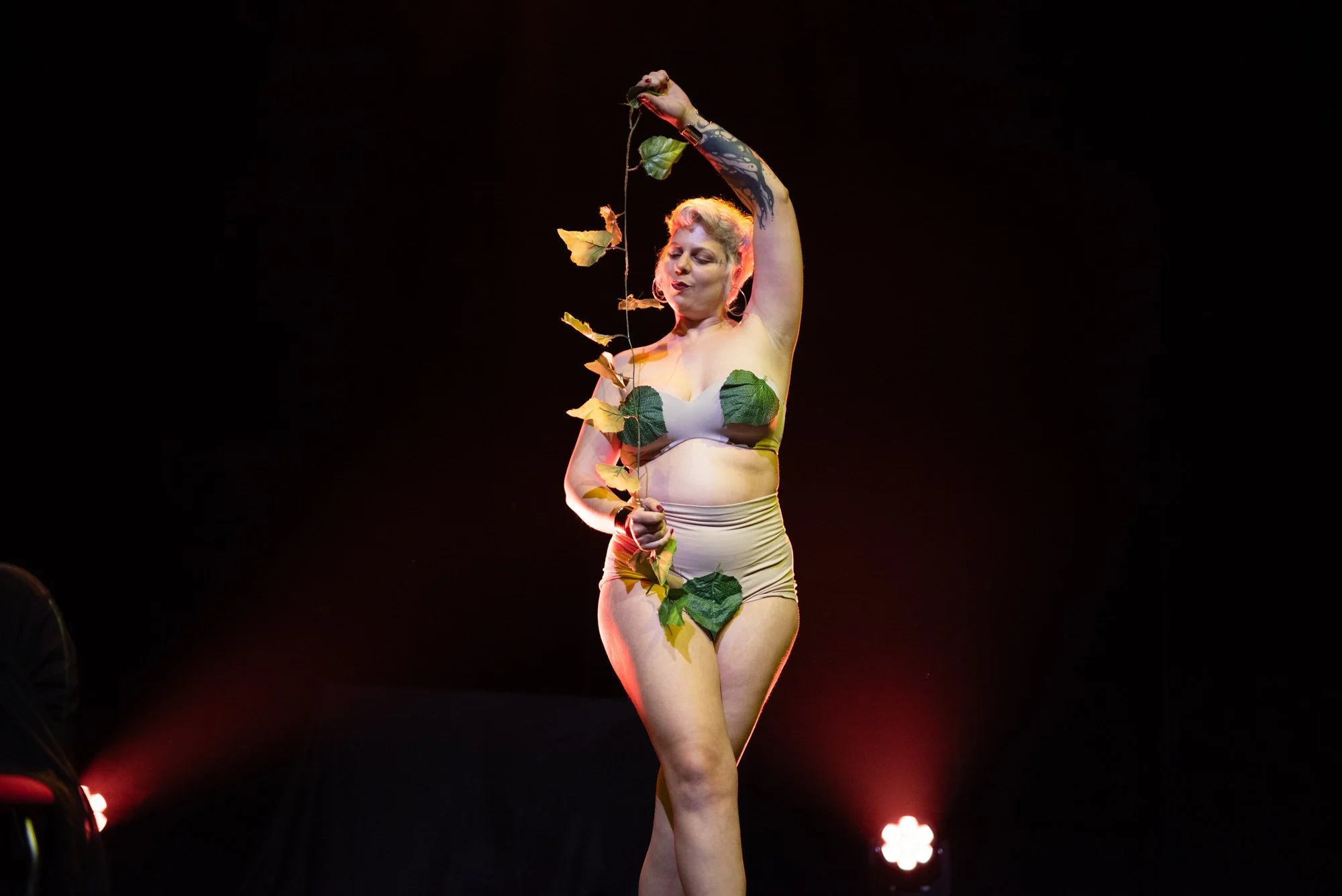 A woman standing on stage, wearing minimal clothing with leaf coverings, holding a long, leafy vine, surrounded by dark background and stage lights.