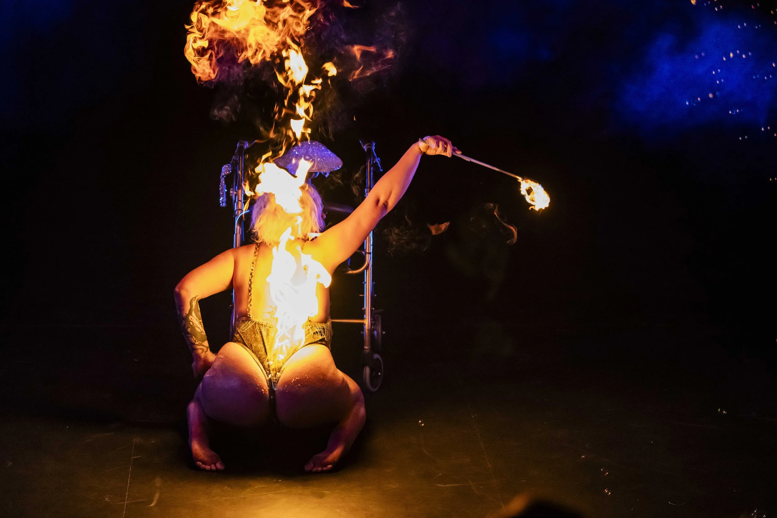 Performer kneeling on the ground, holding a flaming torch in each hand, with a backdrop of dark night sky and bright flames.