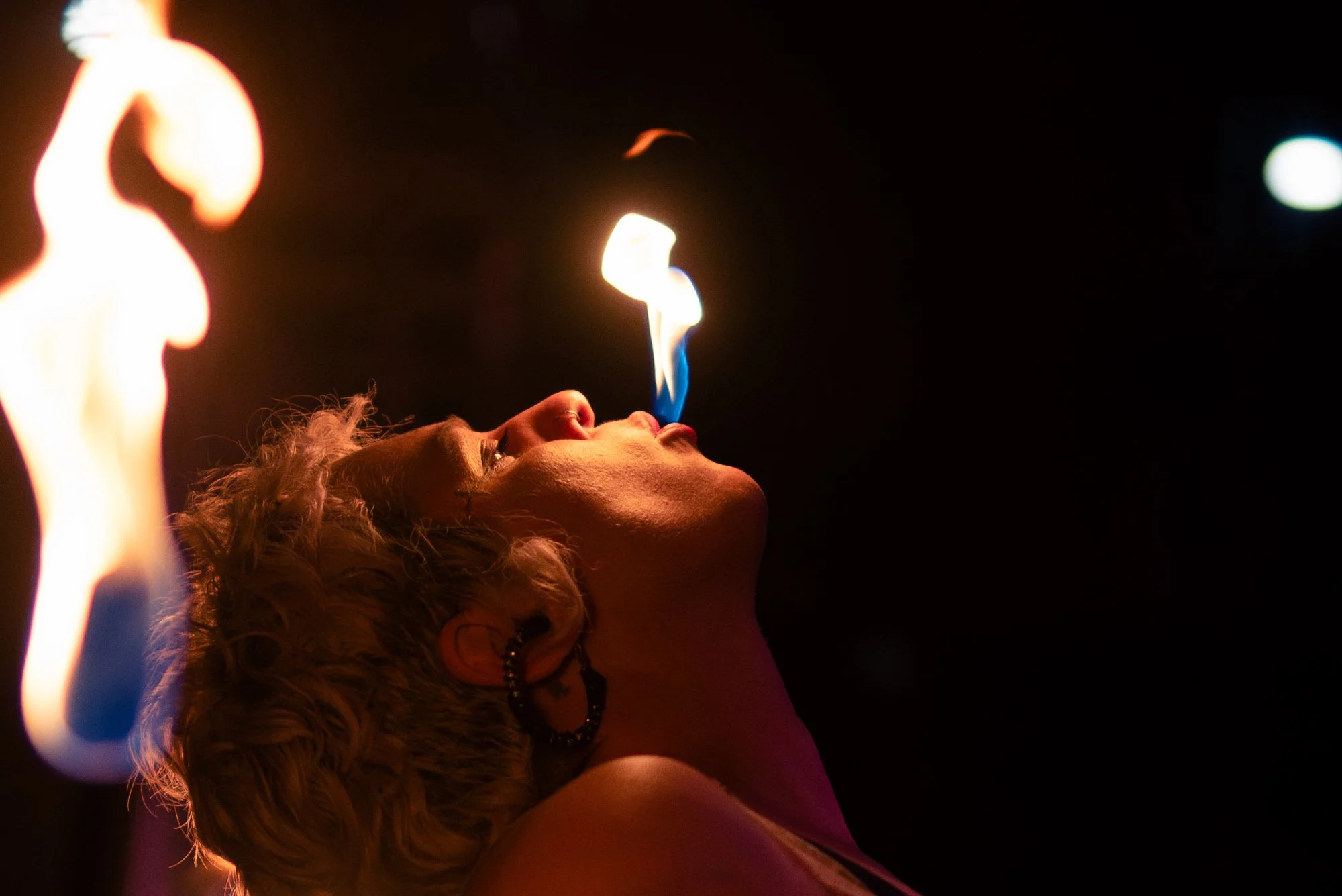 A person with curly hair and earrings blows fire in a dark setting.