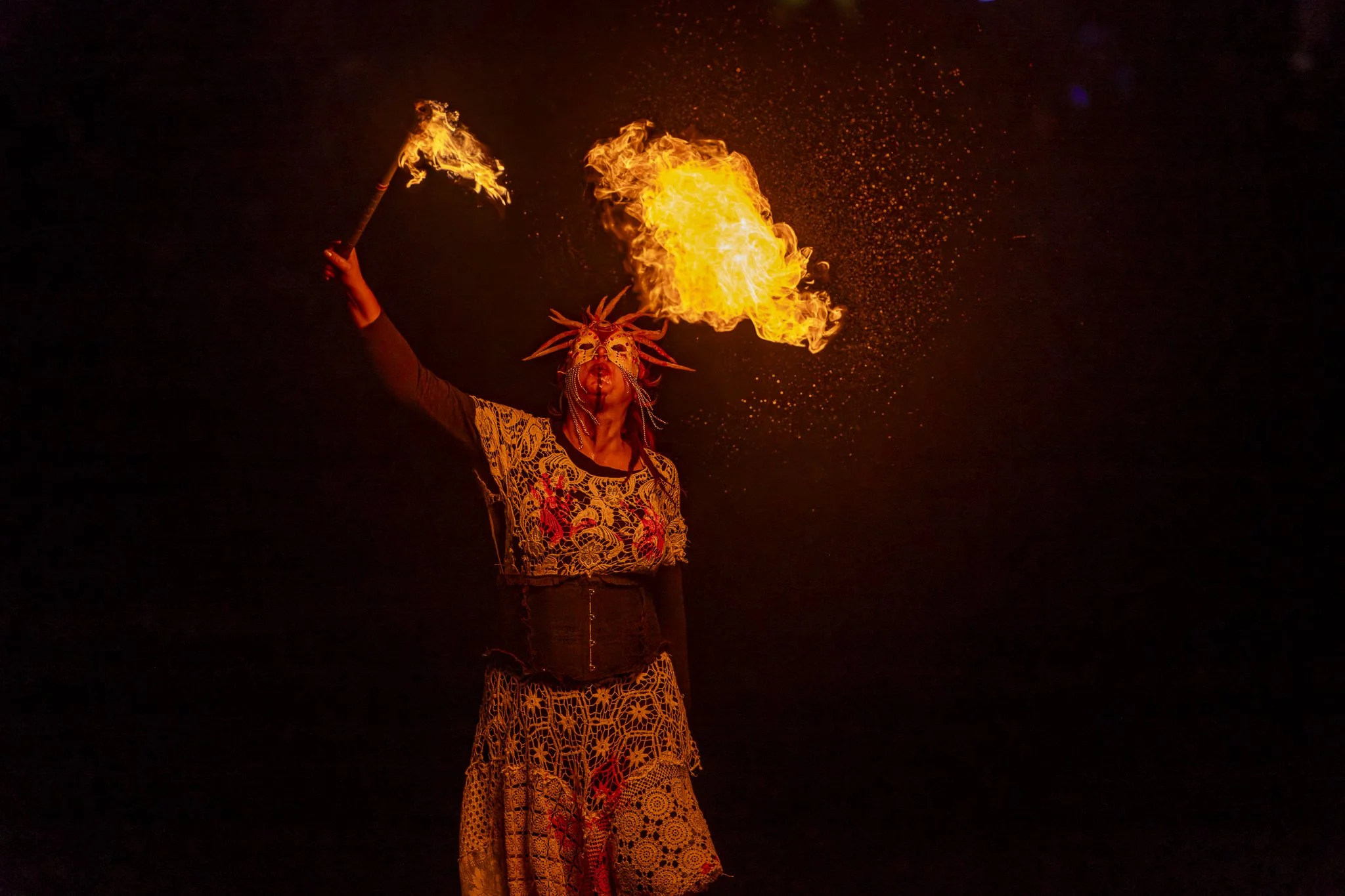 Performer dressed in colorful tribal costume and mask, breathing fire during a night performance.
