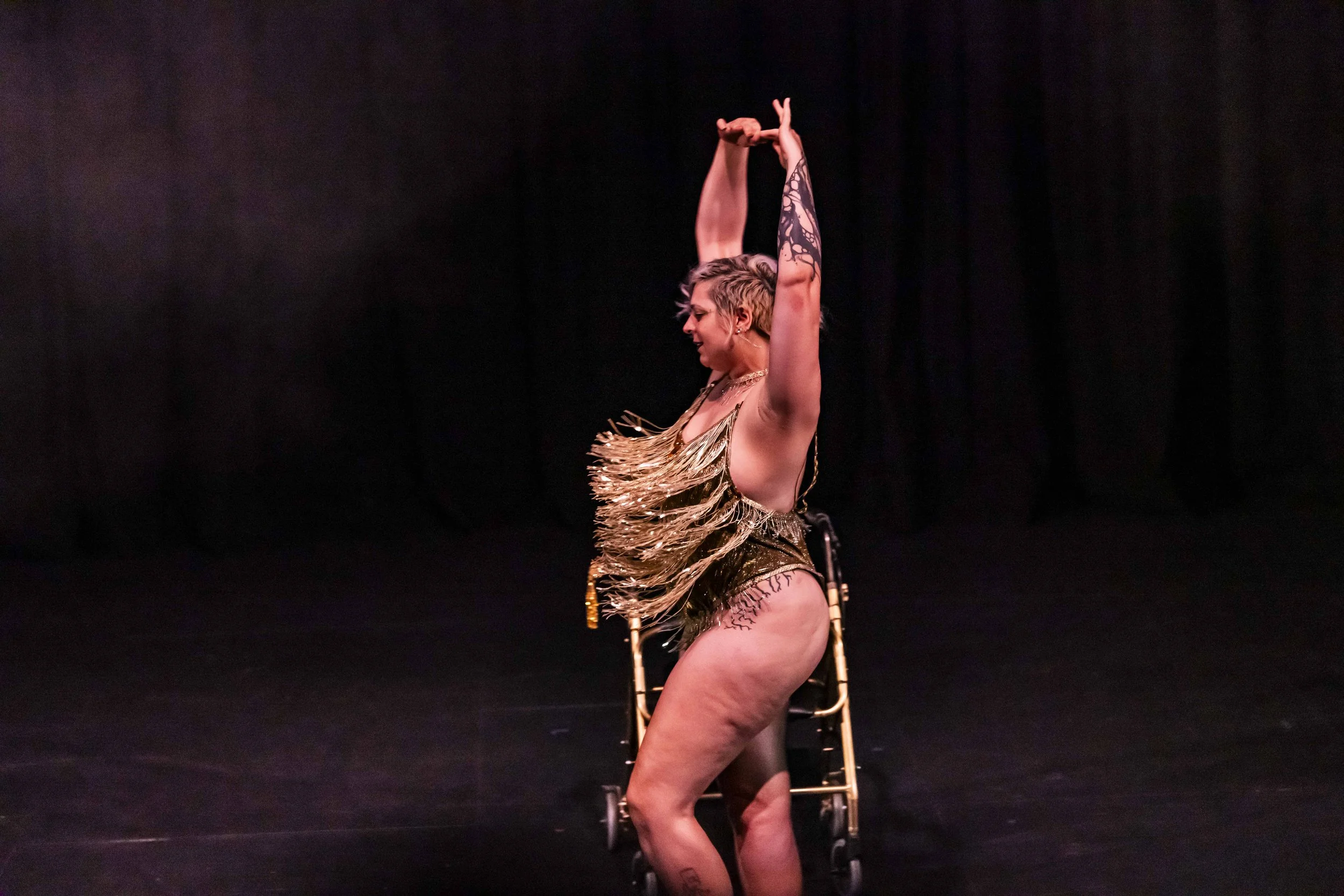 A performer in a gold fringed costume dancing on stage with arms raised, behind a small wheelchair, with a dark background.