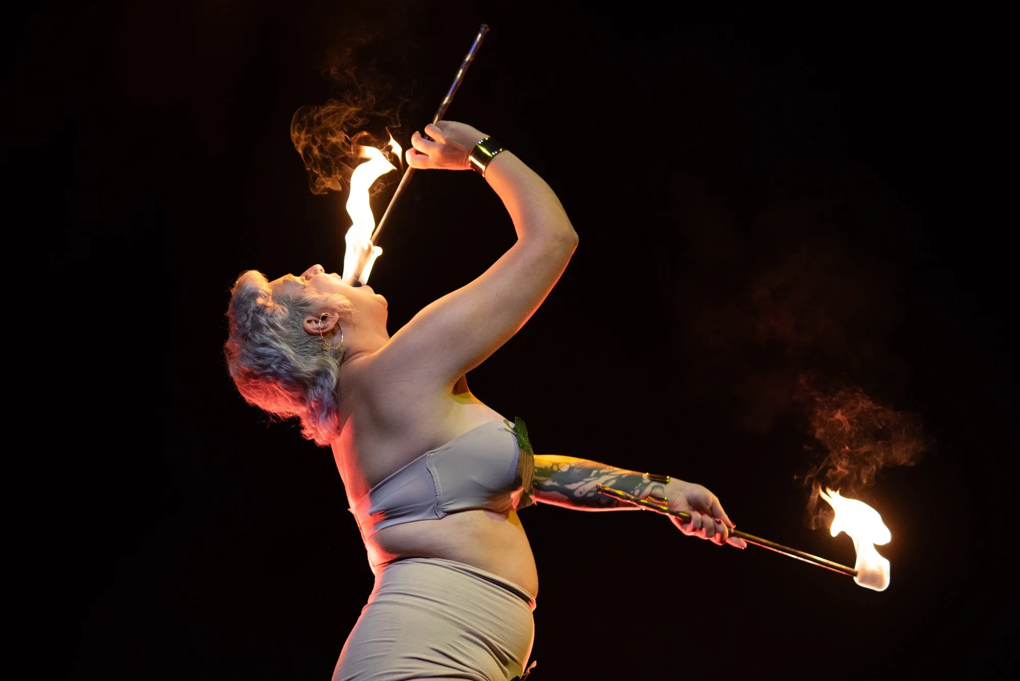 A woman with short, wavy blonde hair performing a fire-eating act, holding a flaming torch close to her mouth in a dark setting.