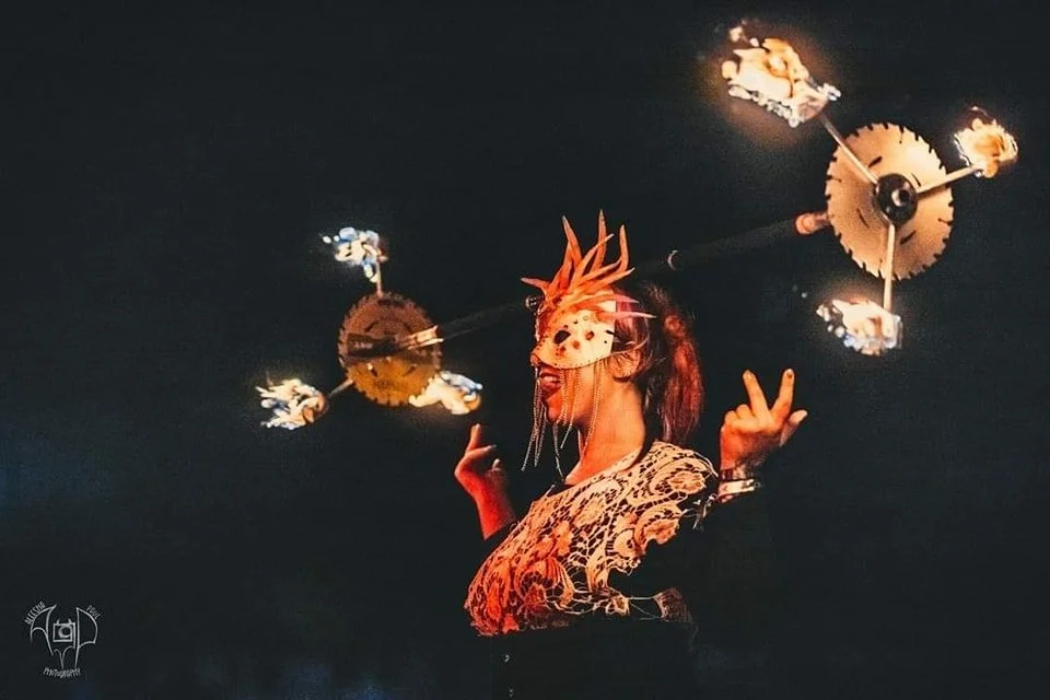 A performer wearing a mask and an elaborate headdress, balancing a flaming object oh her head, performing at night.
