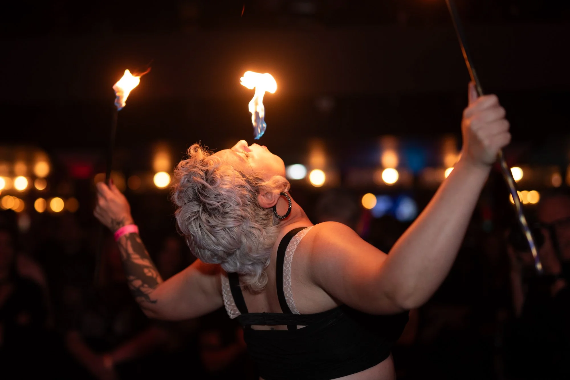 A woman with gray curly hair, wearing a black top and earrings, is performing a fire-breathing act with flames above her mouth at a dimly lit venue.