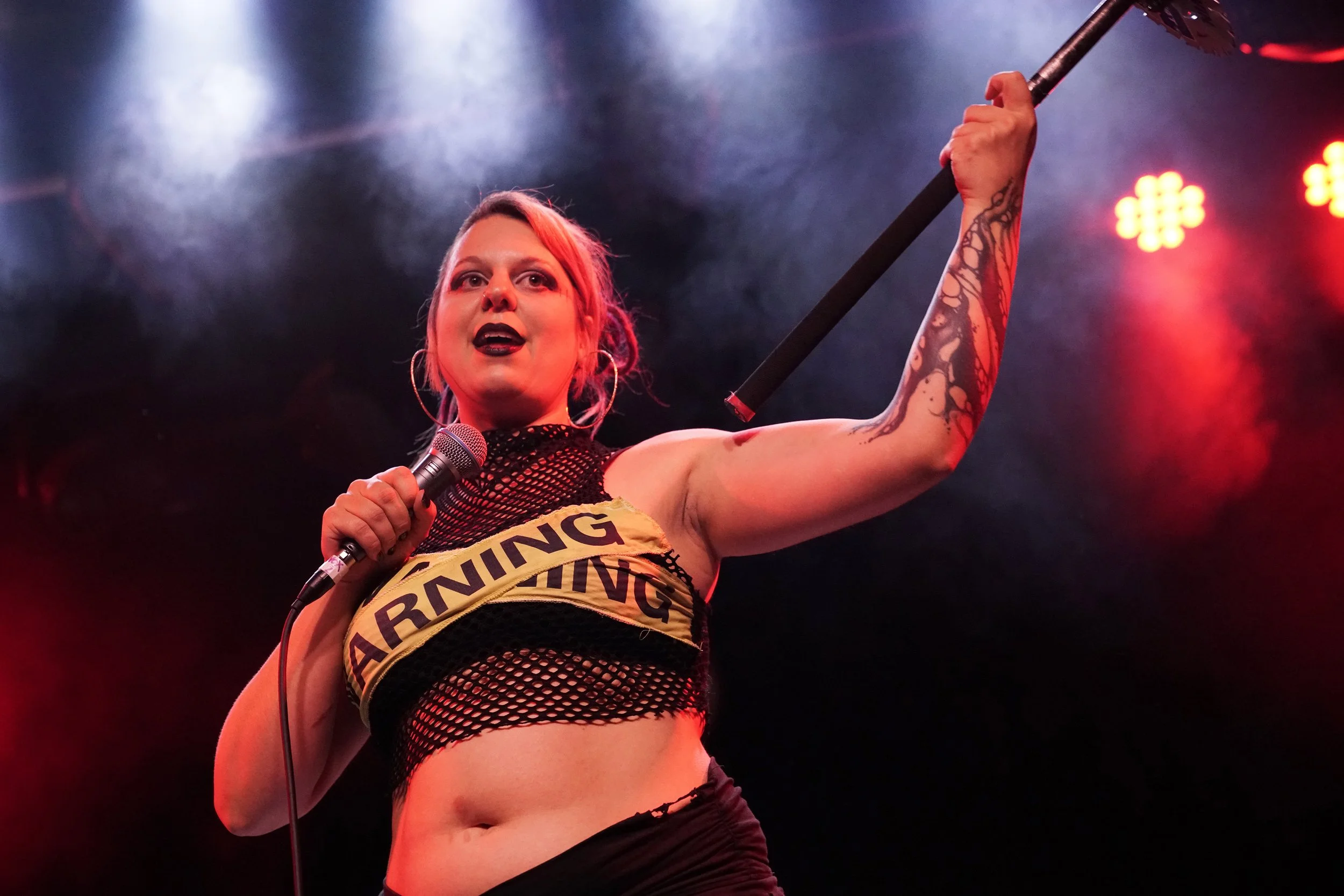 A woman with pink hair and tattoos on her arm is holding a microphone in one hand and an axe over her shoulder on a stage with red and purple lighting.