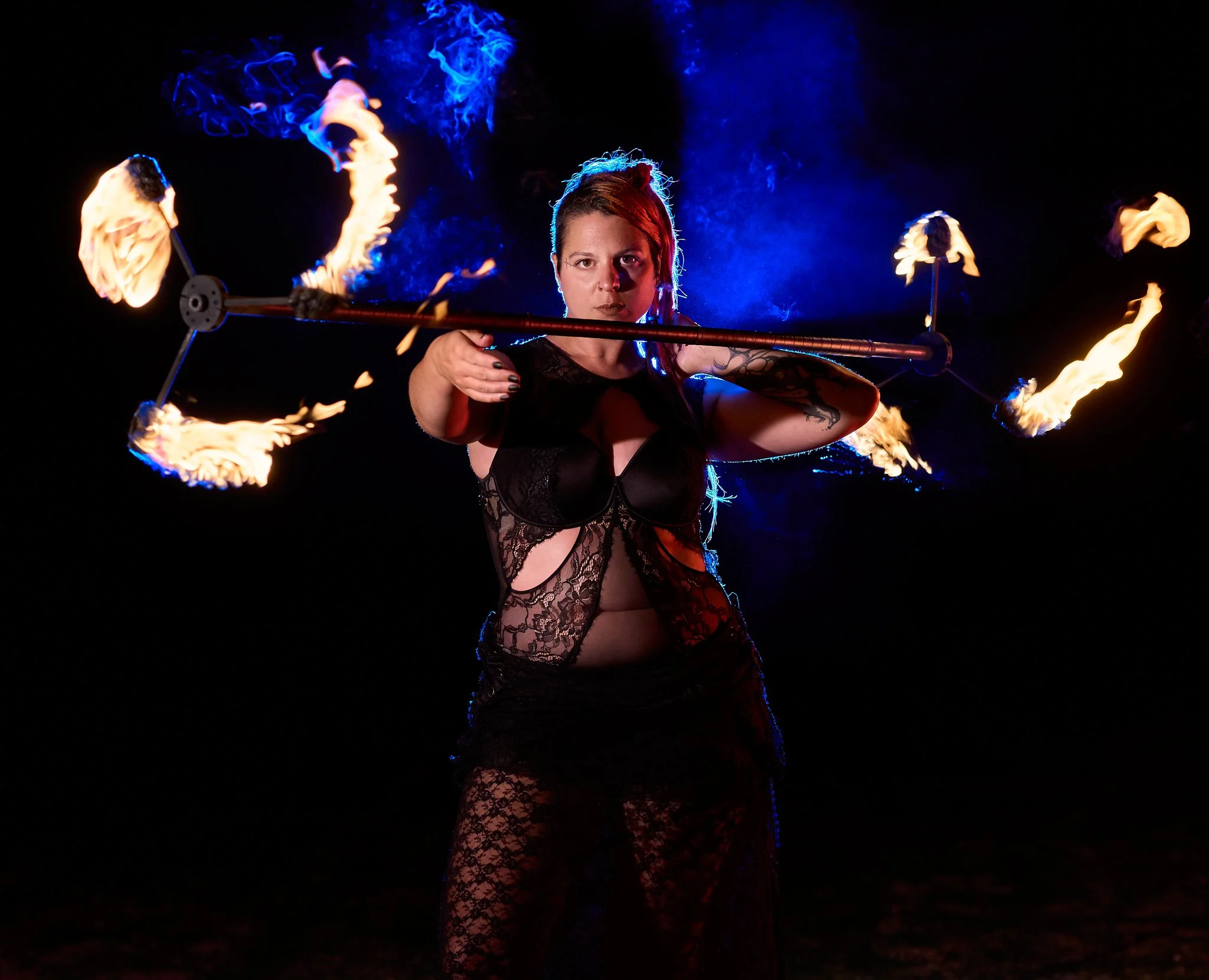 A woman in black lace clothing performs a fire act, spinning a flaming staff in a dark setting with blue smoke behind her.