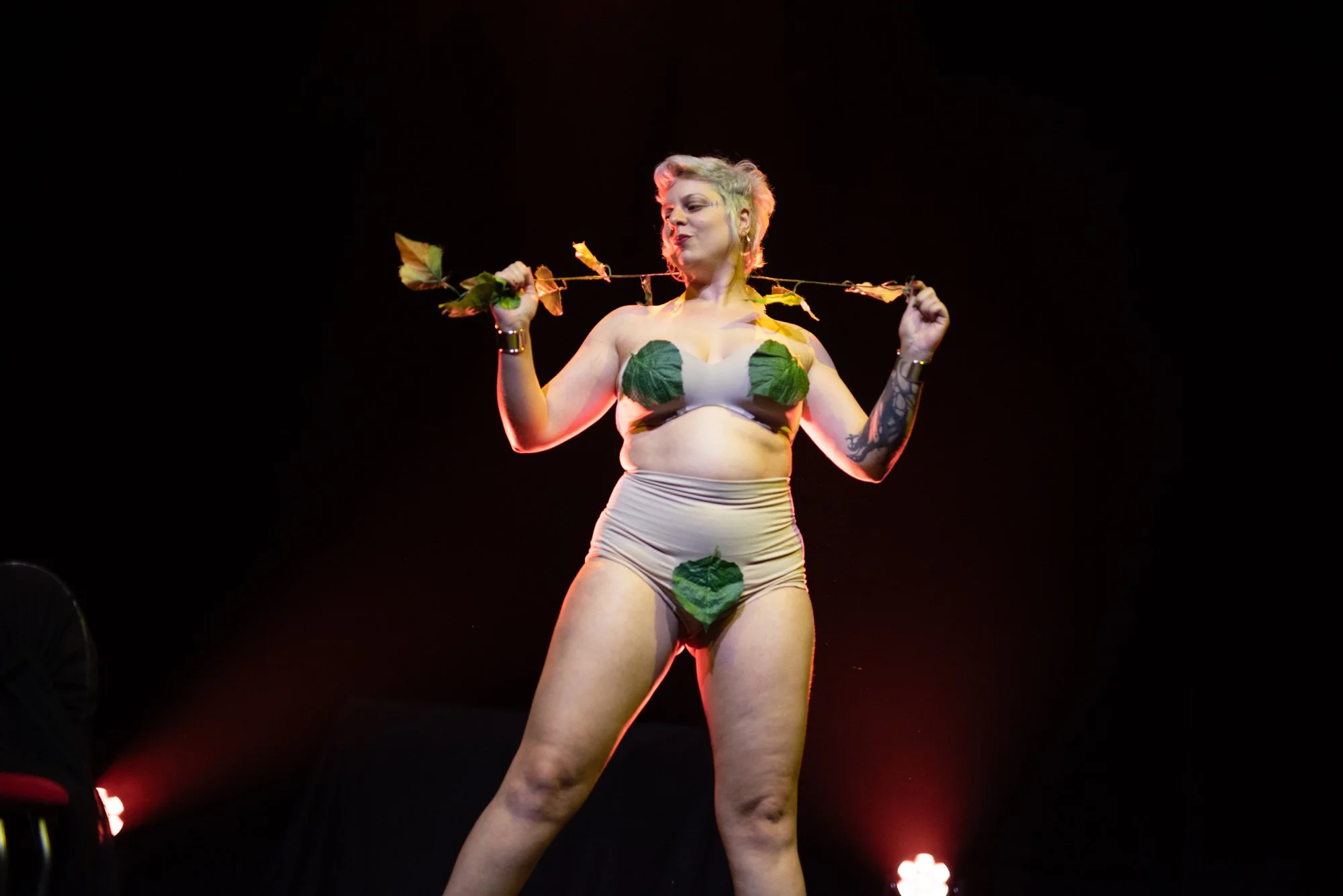 A woman standing on a dark stage, wearing a minimal outfit decorated with green leaves, holding a branch with leaves behind her neck, posing confidently.