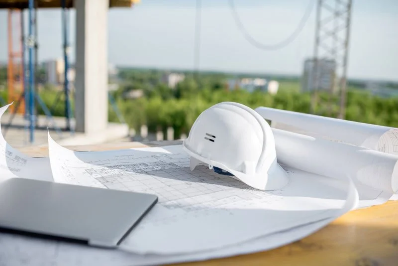 Construction site with blueprints and a white hard hat on a wooden surface, overlooking a green landscape and construction crane in the background.