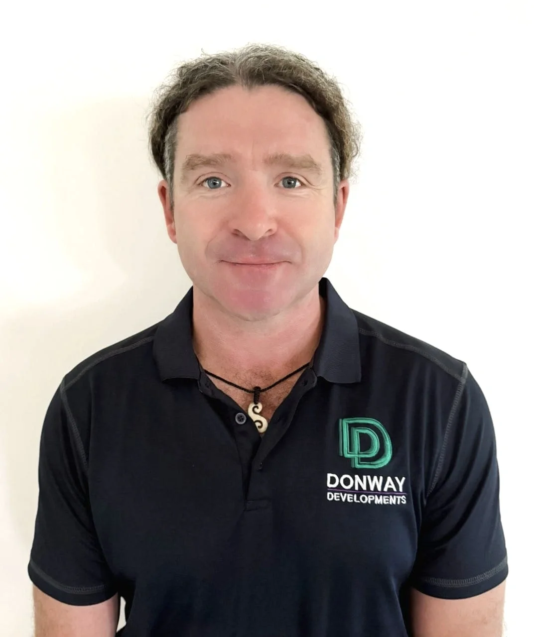 A man with curly brown hair, wearing a black polo shirt with a logo that reads "DONWAY DEVELOPMENTS" and a necklace with a spiral pendant, standing against a plain white background.