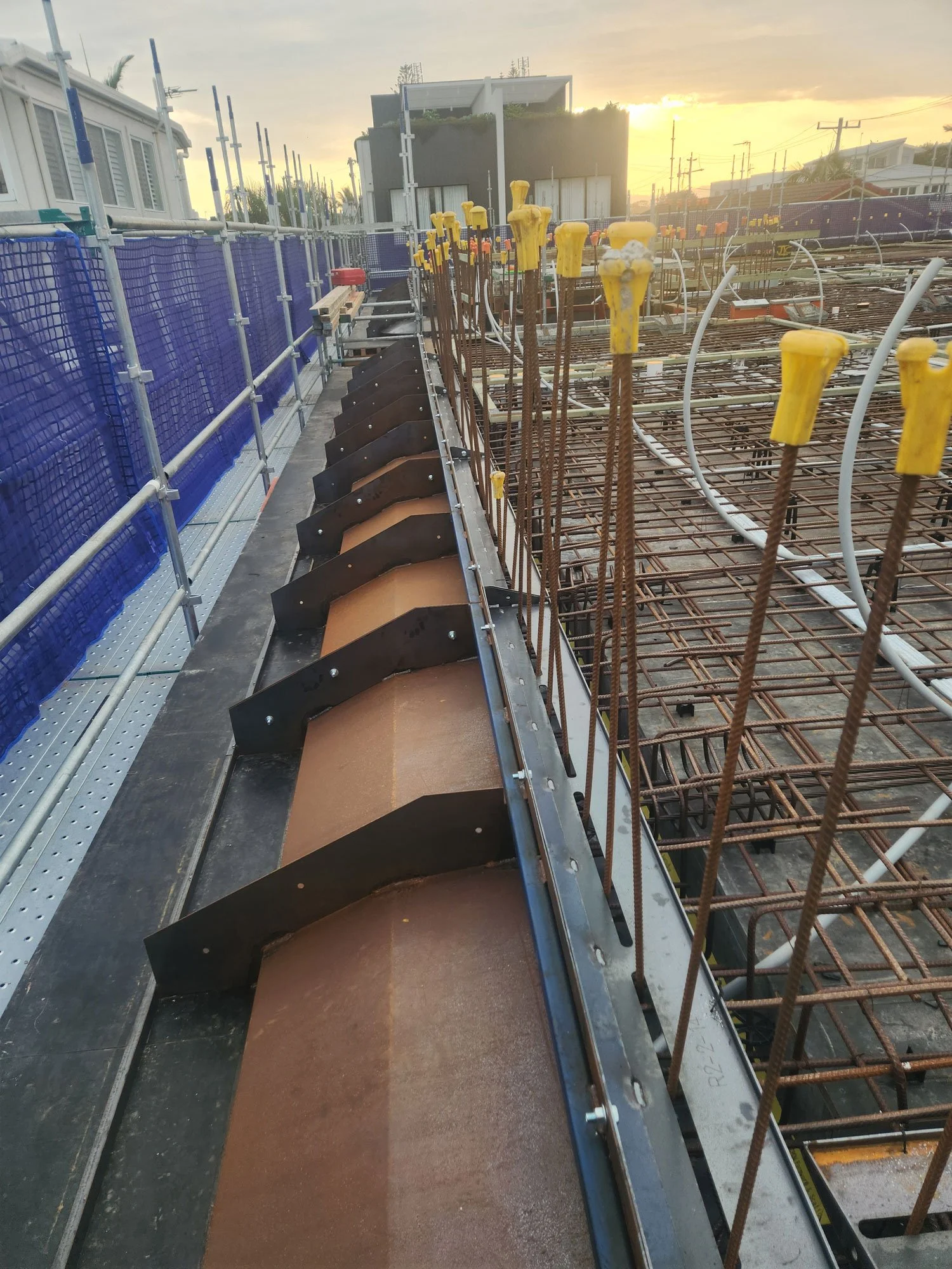 Construction site with steel rebars and yellow plastic caps on top, scaffolding on the left, and a sunset in the background.