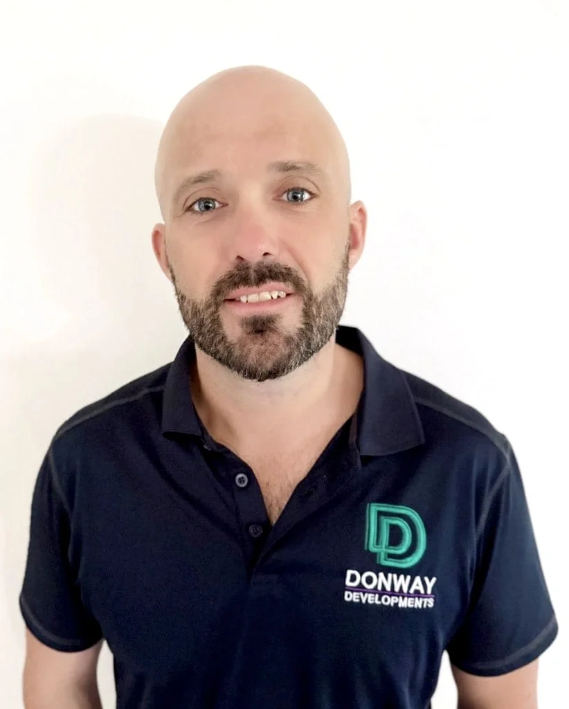 A man with a shaved head and beard wearing a black polo shirt with a logo that says 'Donway Developments' standing against a plain white background.