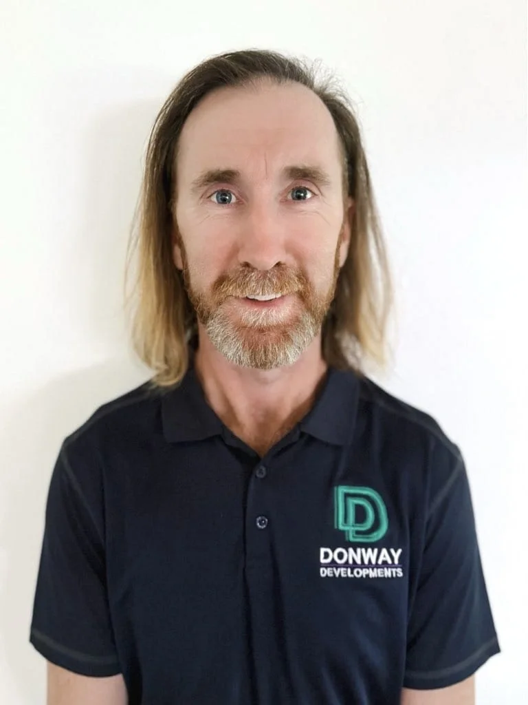 Man with shoulder-length blonde hair and a beard wearing a black collared shirt with 'Donway Developments' logo, standing against a white background.