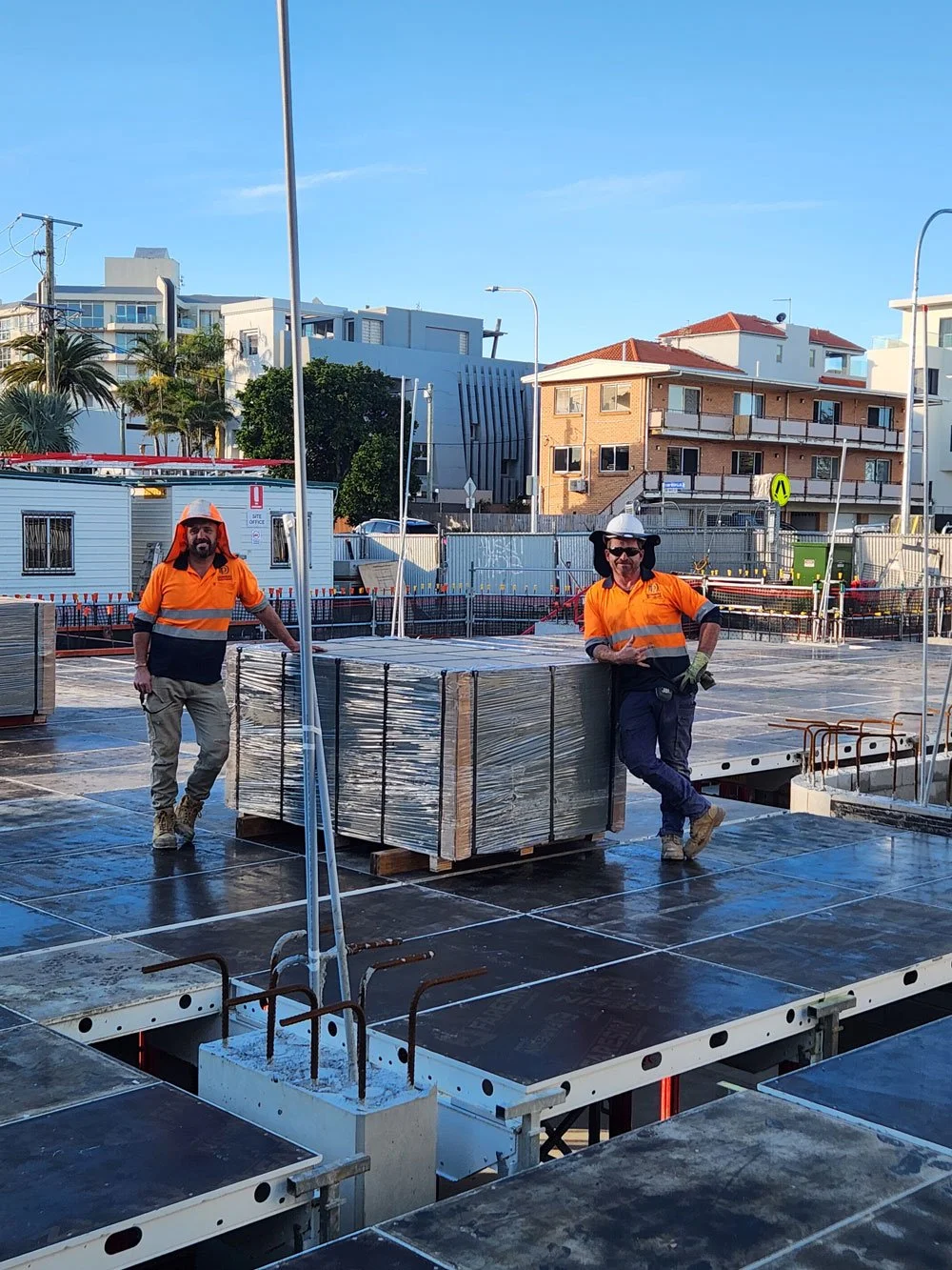 Two construction workers wearing orange shirts and hard hats standing on a construction site with a large pallet of materials, surrounded by buildings and blue sky.