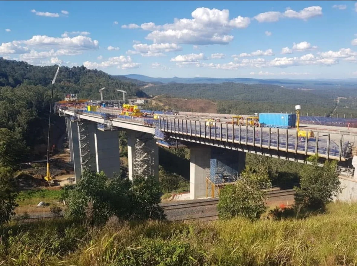 Construction site of a bridge over a river with rolling hills in the background, construction vehicles, and equipment on the bridge.