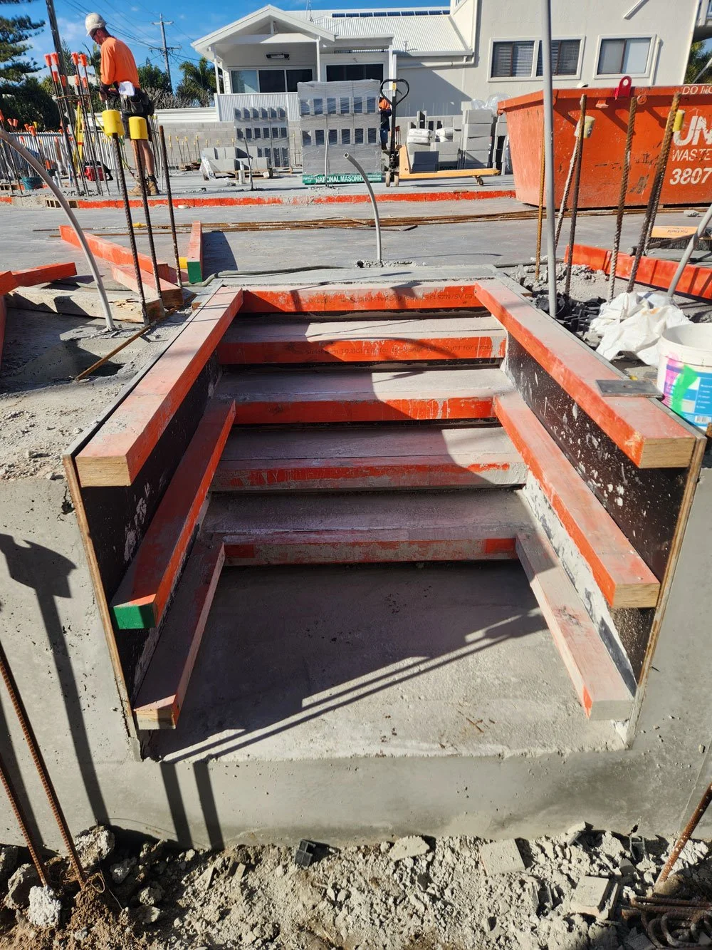 Construction site with framing for stairs being built, surrounded by orange safety barriers, with construction workers and tools in the background.