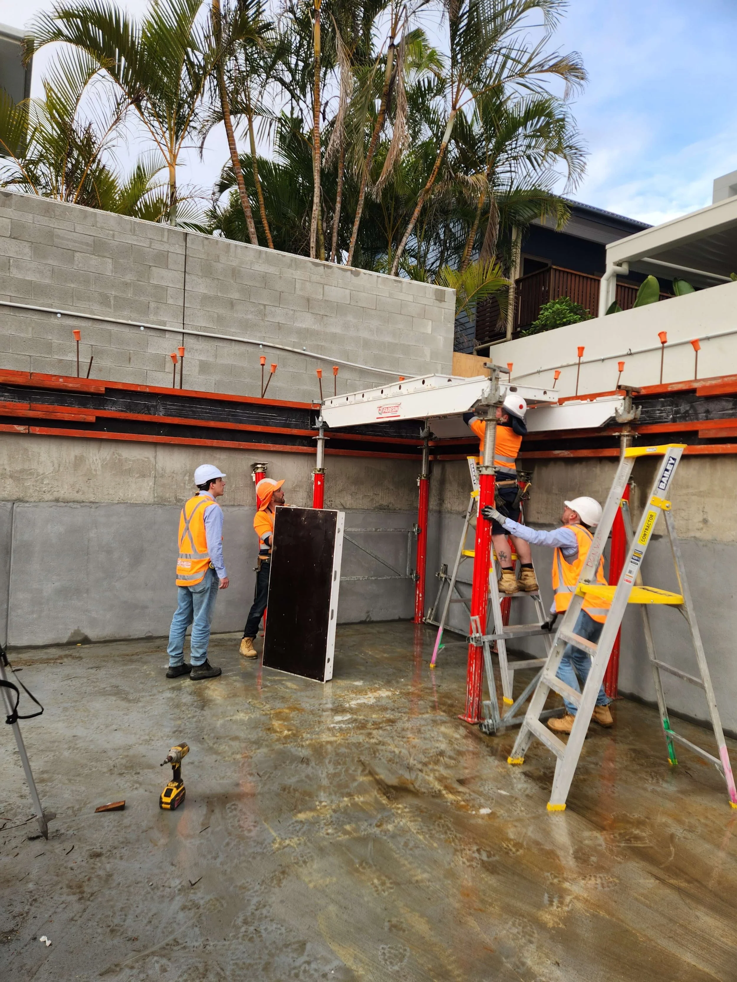 Construction workers wearing hard hats and safety vests working on a building foundation with scaffolding and a concrete wall, with trees in the background.